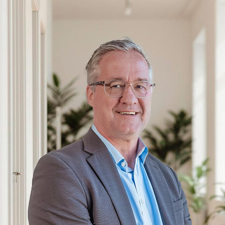 Mann im Anzug steht in einem hellen Flur mit Pflanzen im Hintergrund.