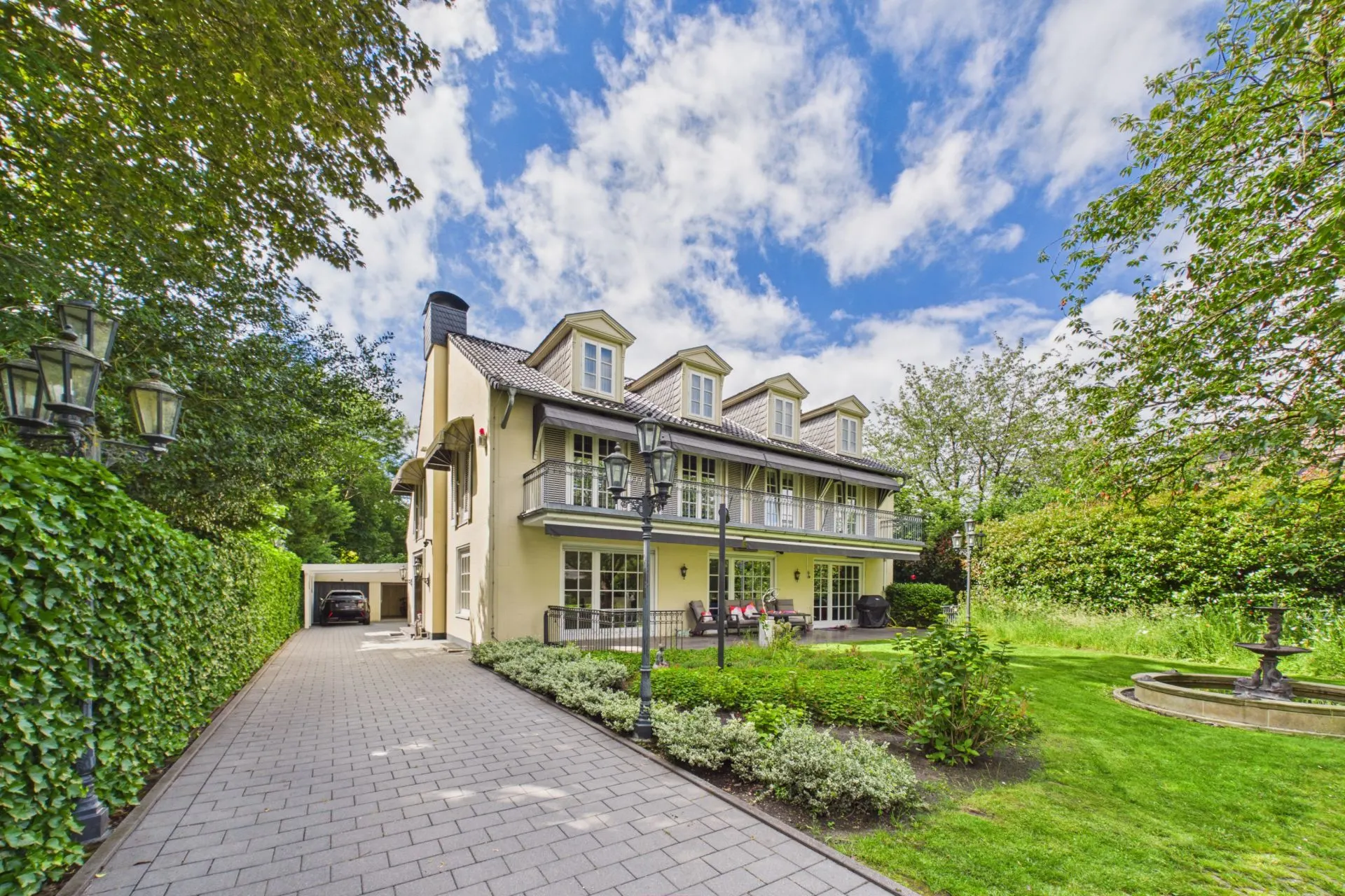 Großes Wohnhaus mit Garten, Auffahrt und Springbrunnen bei blauem Himmel.