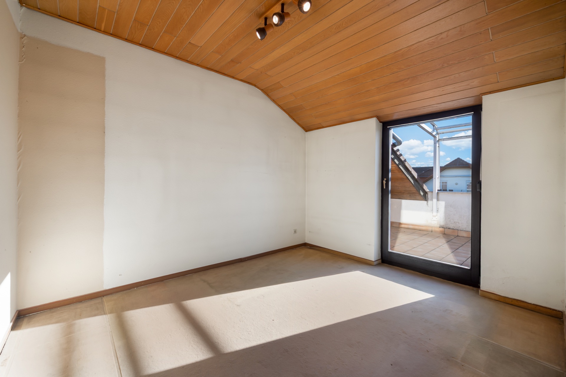 Leeres Zimmer mit Holzdecke, großer Glastür und Zugang zu einem Balkon.