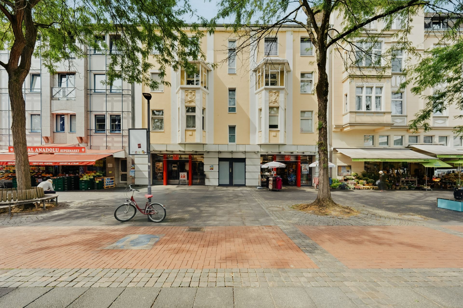 Mehrstöckiges Wohnhaus mit Bäumen, Ladenlokalen und einem Fahrrad vor dem Gebäude.