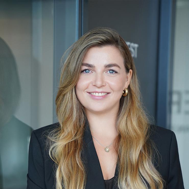 Frau mit langen blonden Haaren und schwarzem Blazer steht vor Glaswand.