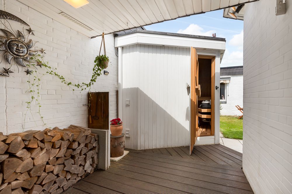 Überdachte Terrasse mit Holzstapeln, einer Pflanze und offenem Zugang zu einem kleinen weißen Raum.