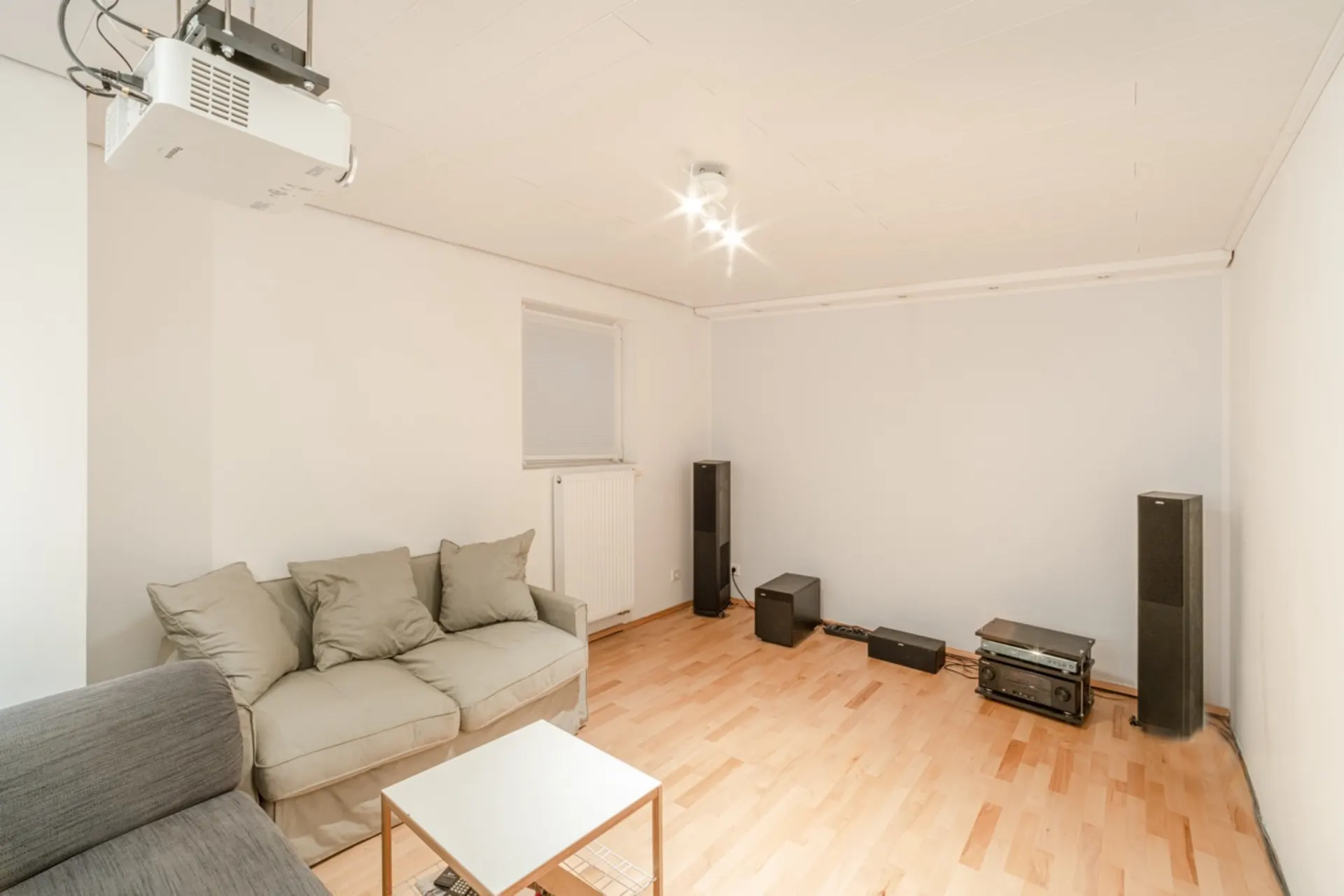 Helles Wohnzimmer mit Sofa, Couchtisch, Lautsprechern und einem Beamer an der Decke.
