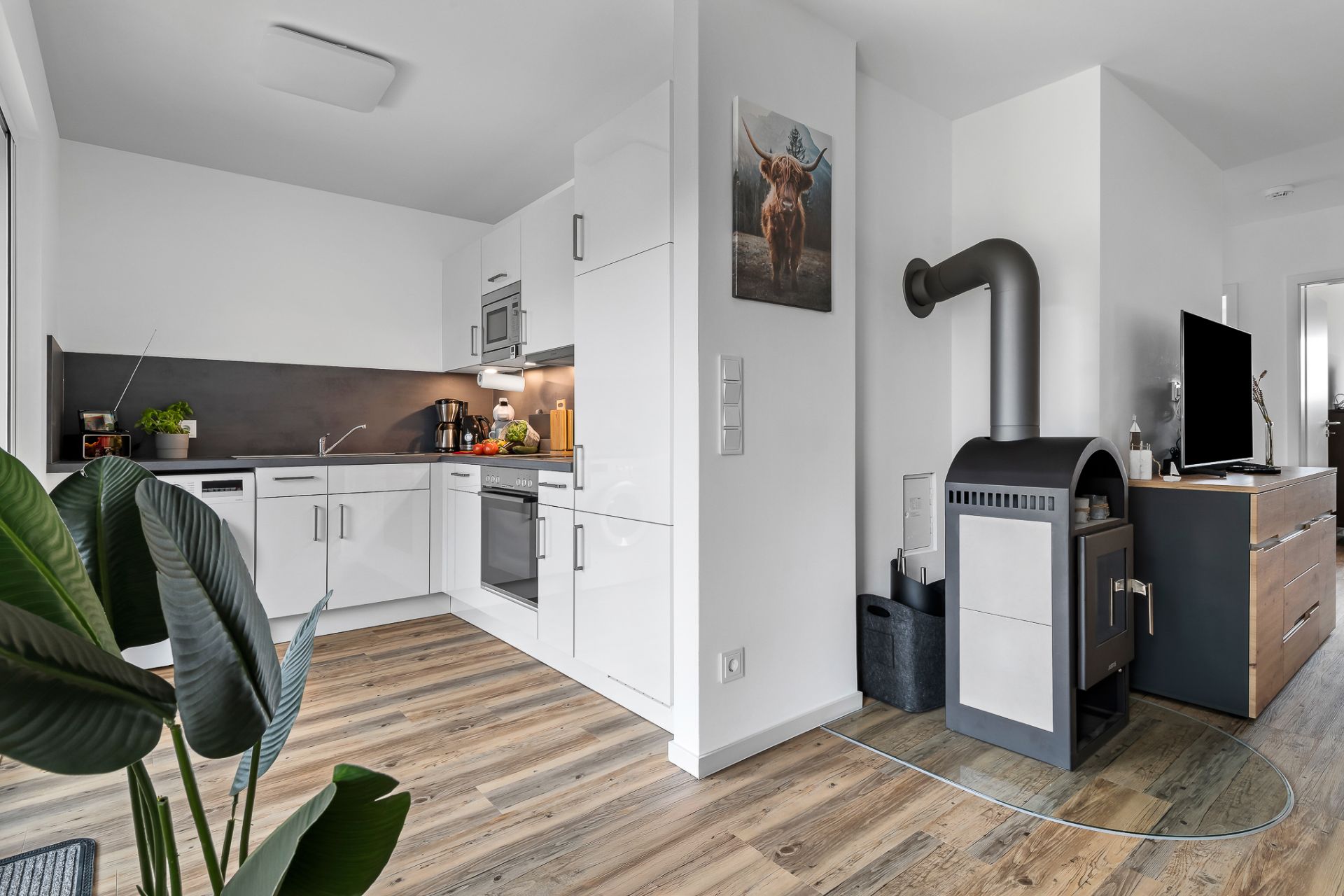 Moderne Einbauküche mit weißen Schränken, Holzofen und Pflanzen auf Holzfußboden.
