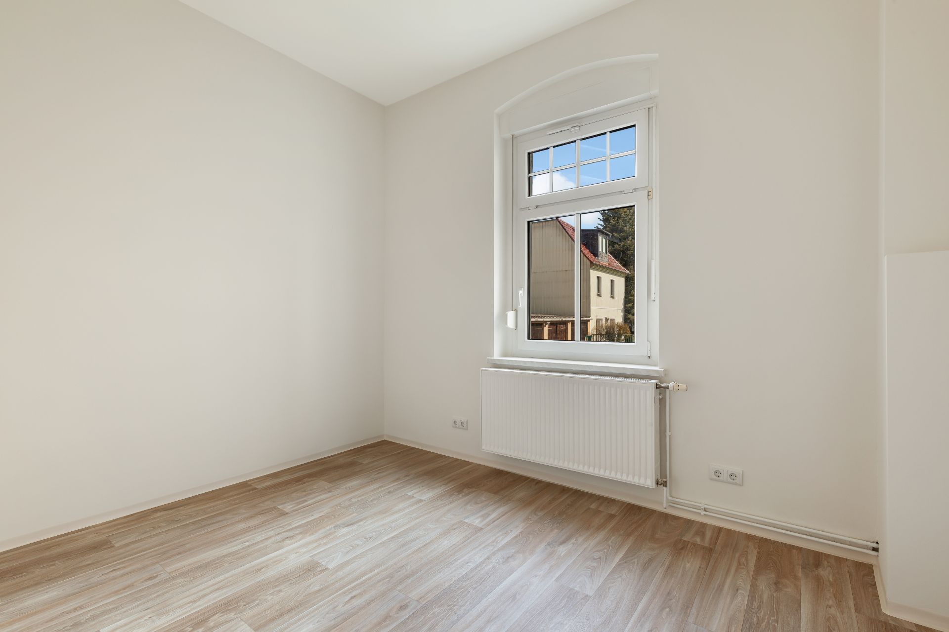 Helles leeres Zimmer mit Fenster, Heizkörper und hellem Holzfußboden.