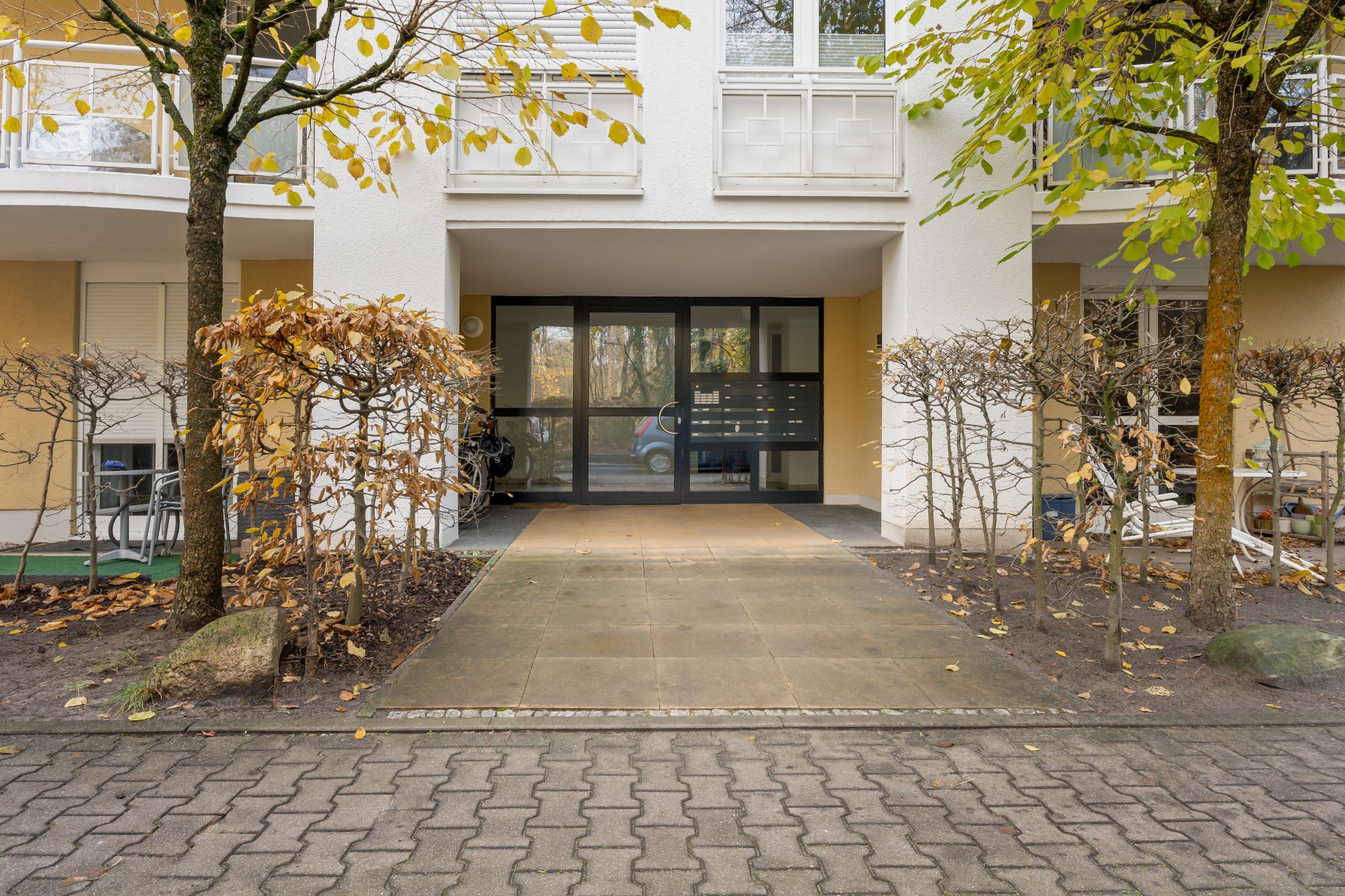 Eingang eines Wohnhauses mit Glasfront und Briefkästen, umgeben von Bäumen und niedrigen Sträuchern.