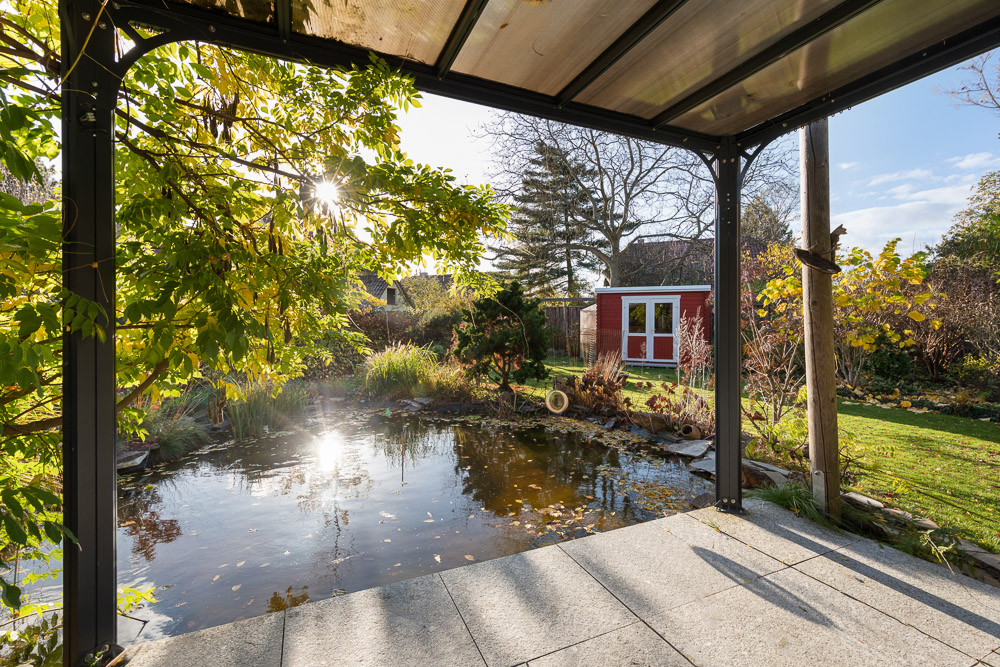 Blick von überdachter Terrasse auf einen Gartenteich mit grüner Bepflanzung und rotem Gartenhaus.