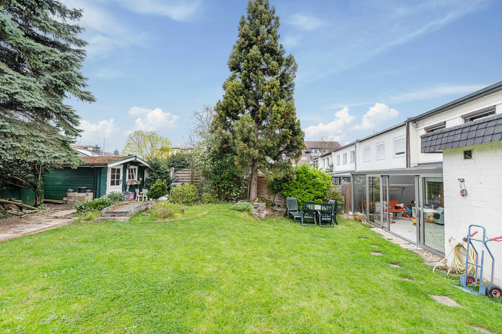 Grüner Garten mit Terrasse, großem Baum, Gartenhaus und Sitzgruppe hinter einem Wohnhaus.
