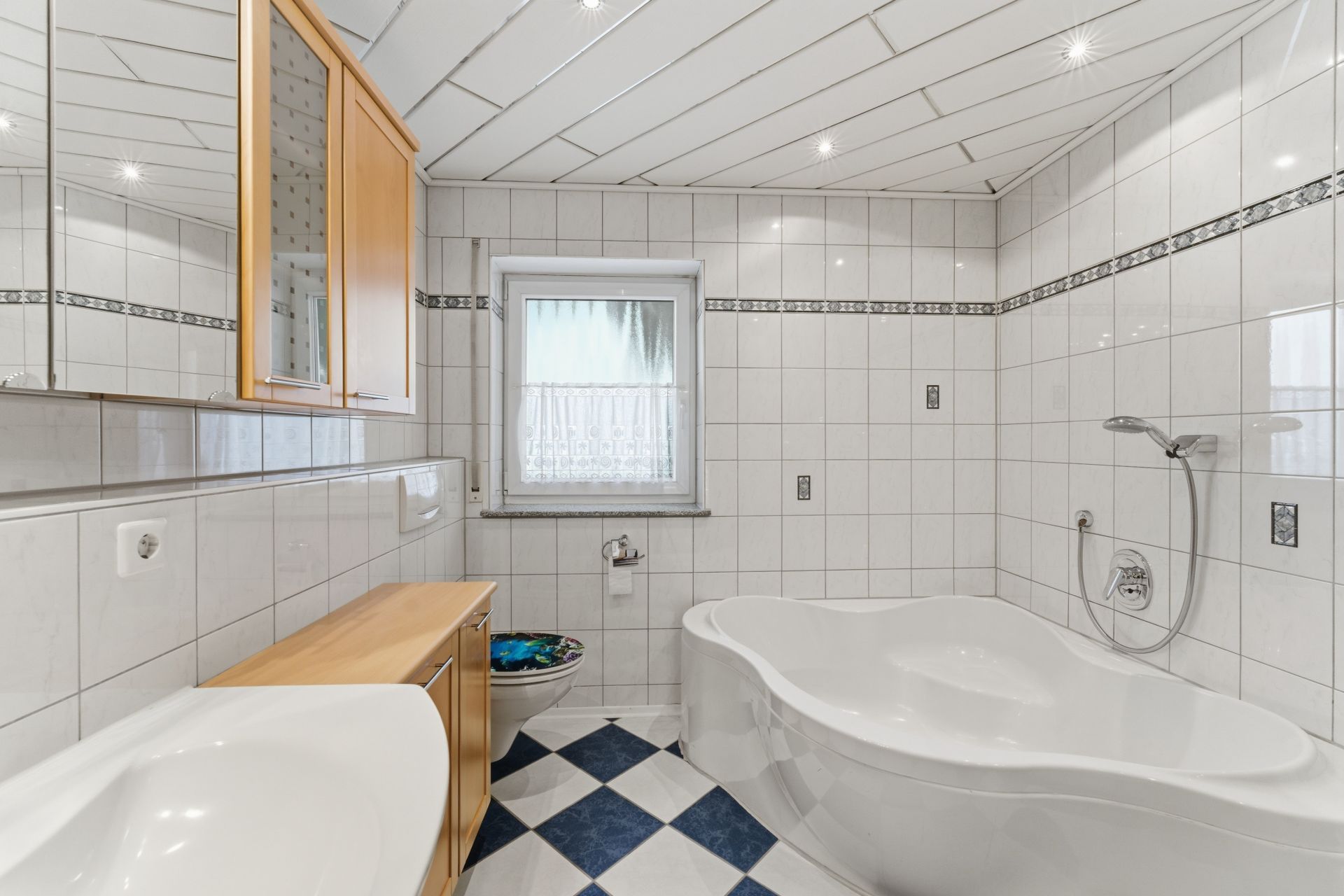 Modernes Badezimmer mit Eckbadewanne, Waschbecken, Toilette und Fenster, weiße Fliesen an Wänden und Boden.