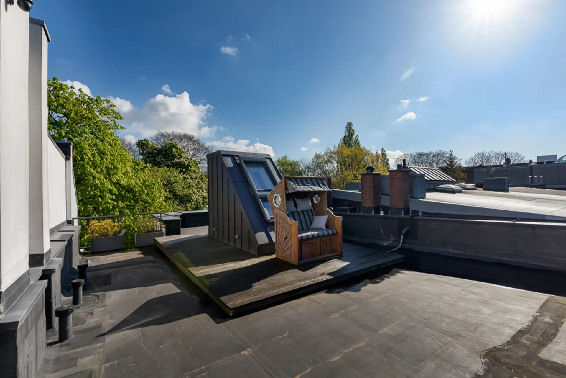 Dachterrasse mit Strandkorb, Holzboden und Blick auf Bäume, bei sonnigem Wetter.