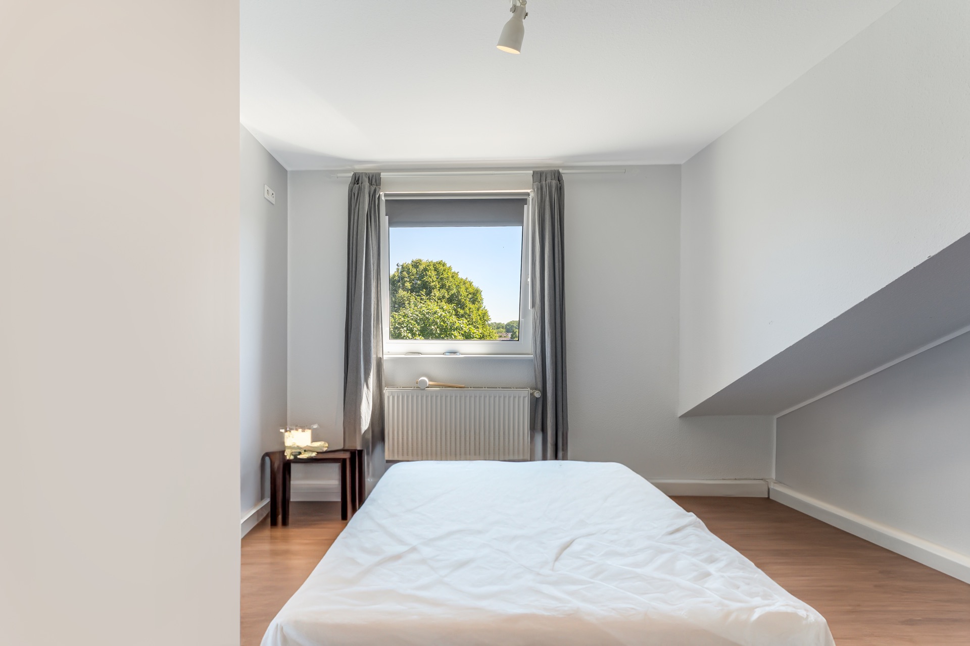 Helles Schlafzimmer mit niedrigem Bett, Fenster, grauen Vorhängen und kleinem Tisch mit Kerzen.