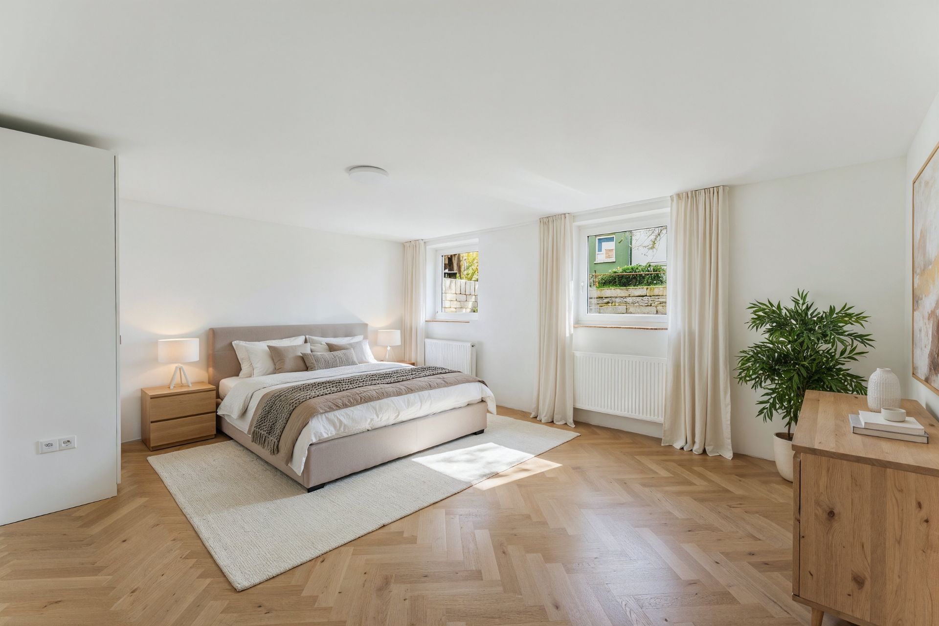 Helles Schlafzimmer mit großem Bett, zwei Nachttischen, Pflanze und Kommode auf Parkettboden.