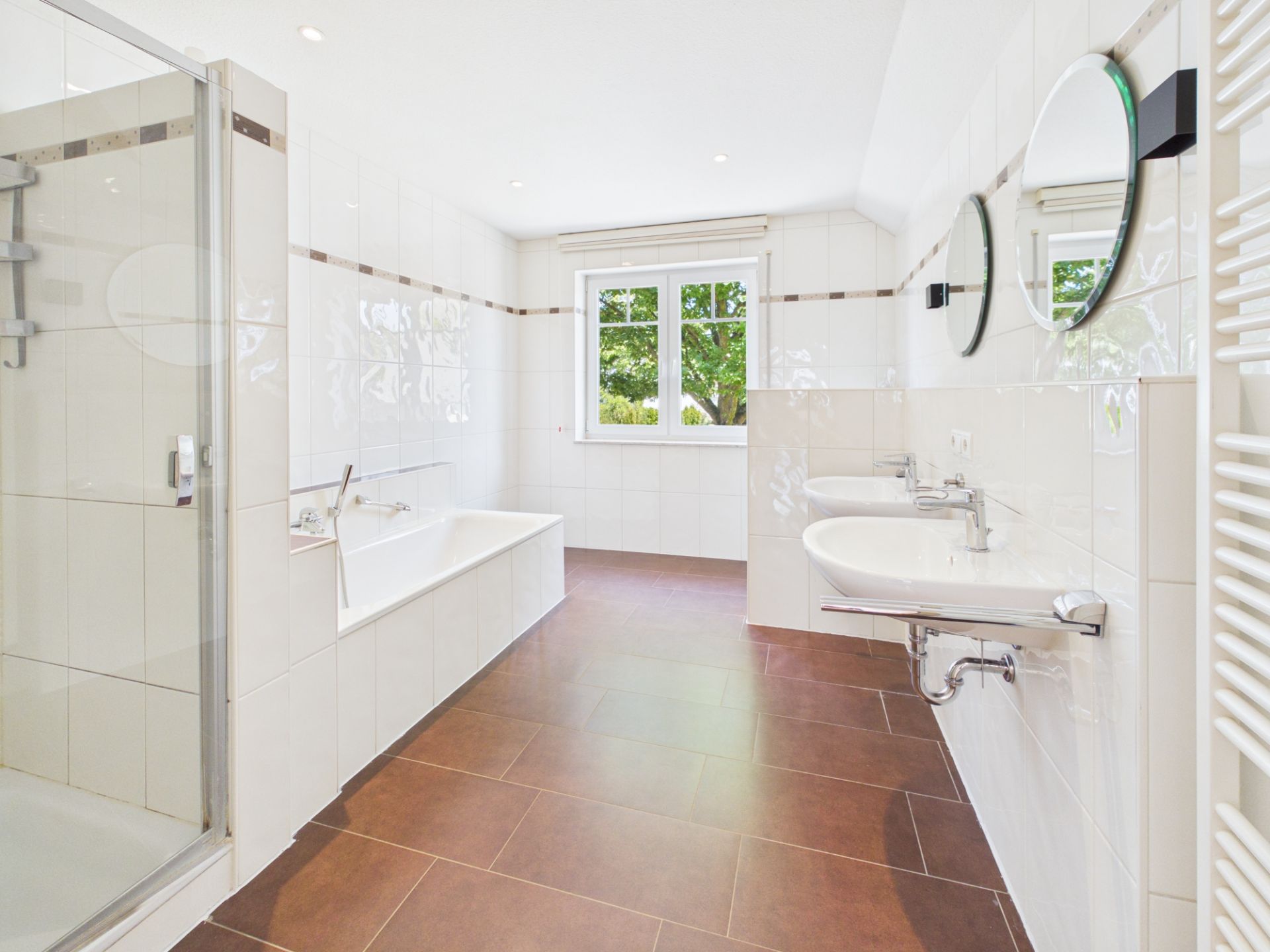 Helles Badezimmer mit Dusche, Badewanne, zwei Waschbecken und Fenster, brauner Bodenfliese.