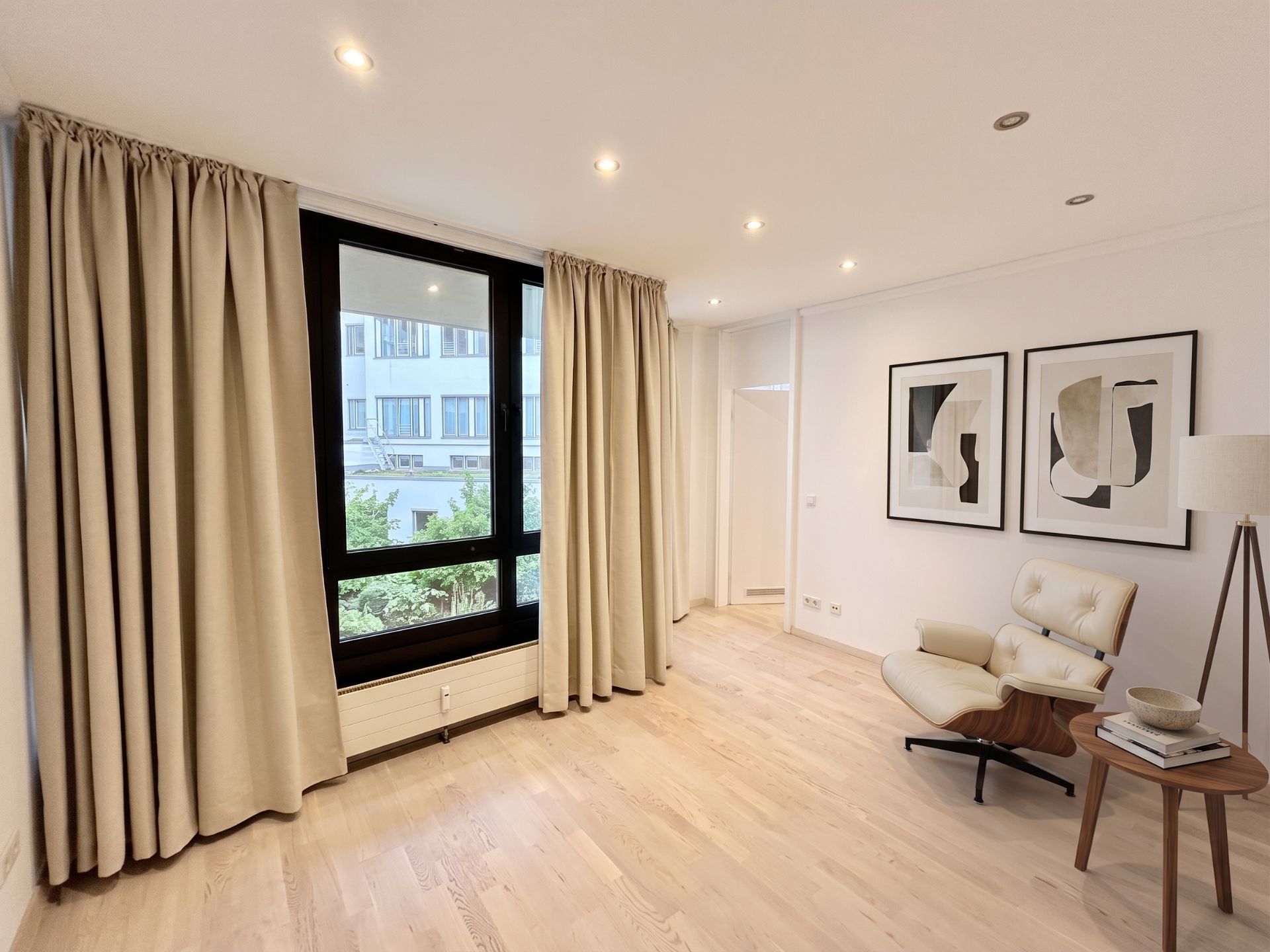 Helles Wohnzimmer mit großem Fenster, beigen Vorhängen, Sessel und zwei abstrakten Bildern an der Wand.