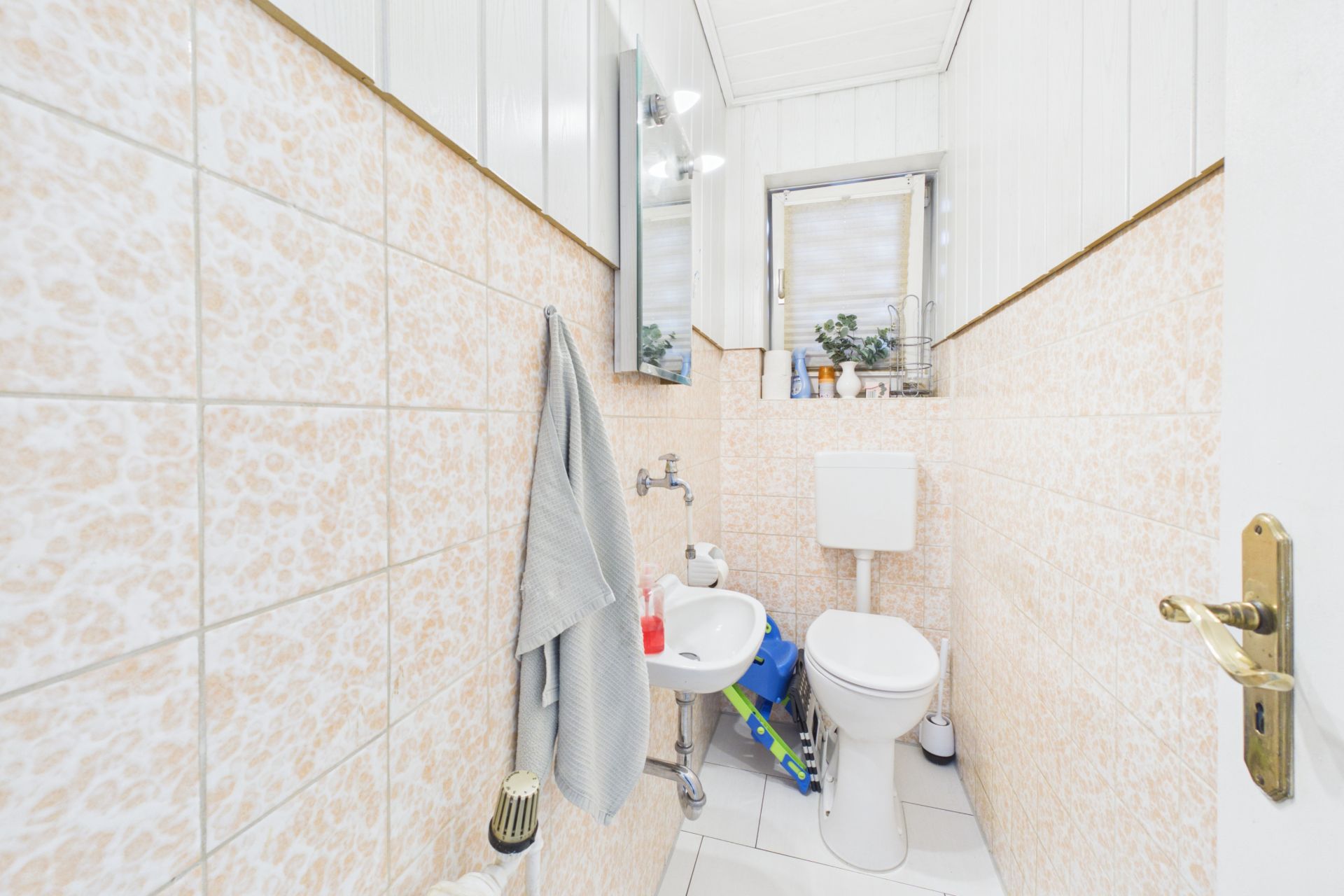 Kleines Bad mit Toilette, Waschbecken, Handtuch, Spiegel und Fenster mit Pflanzen.