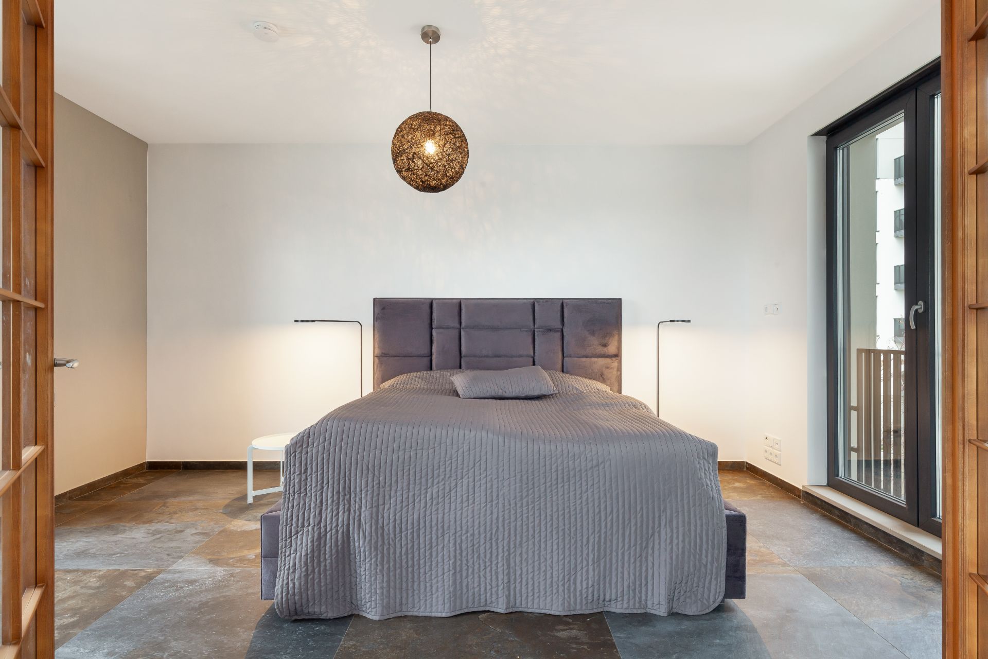Modernes Schlafzimmer mit grauem Bett, zwei Stehlampen und großem Fenster.