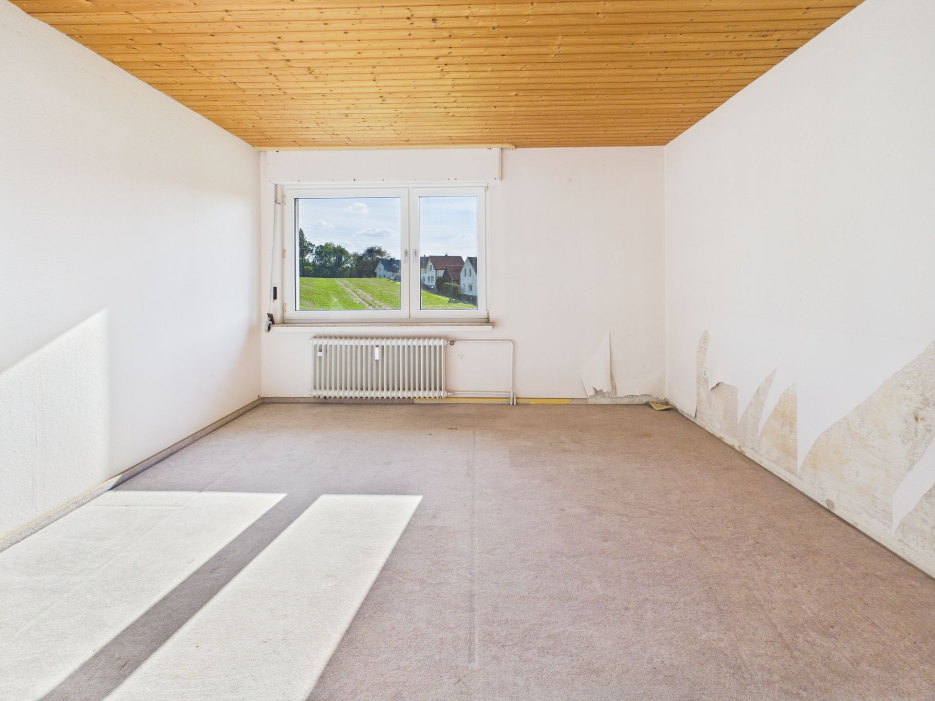 Leeres Zimmer mit Holzdecke, großem Fenster, Heizung und teilweise abgelöster Tapete an den Wänden.