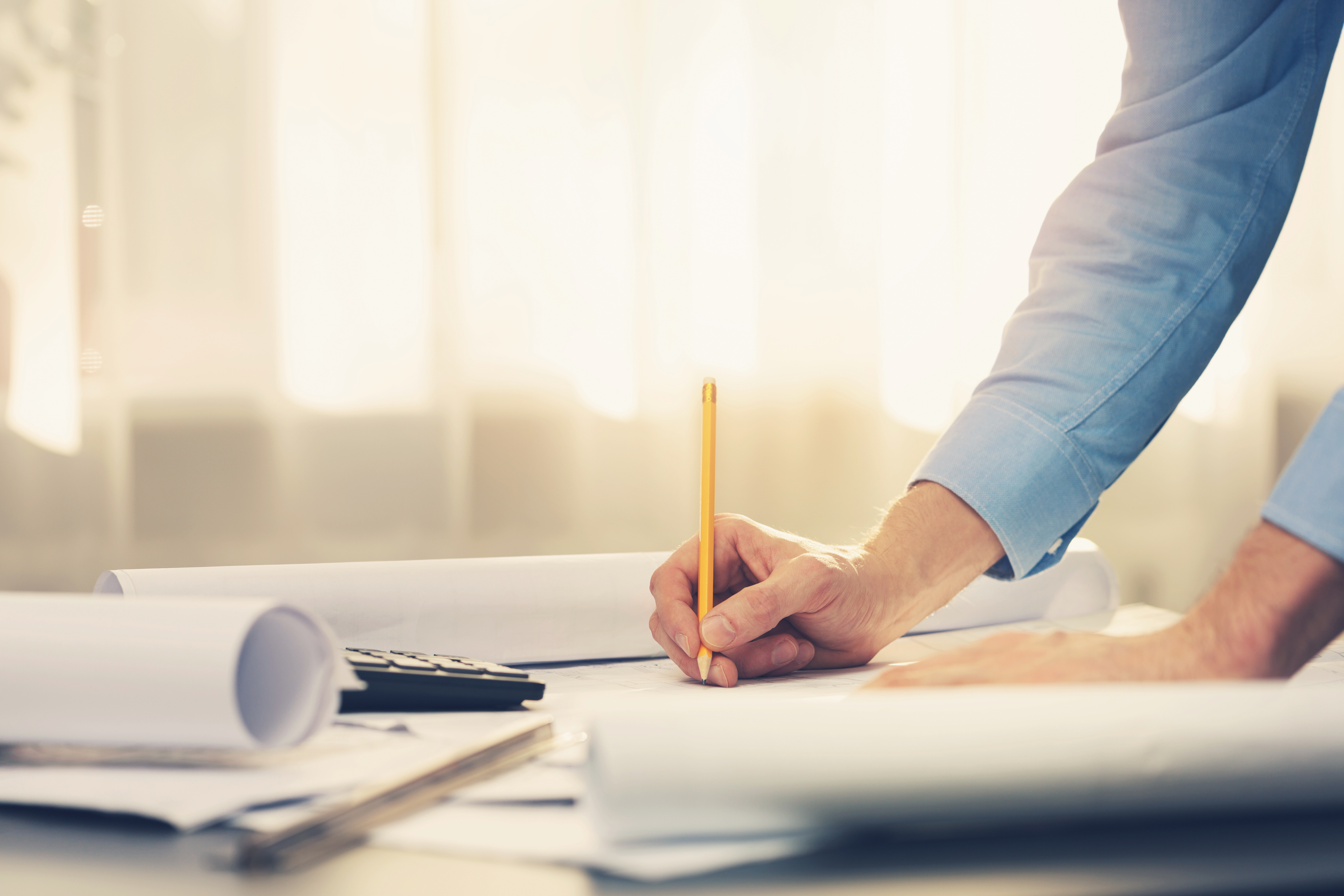 Mann schreibt mit Bleistift auf Papier neben Plänen und Taschenrechner auf Schreibtisch.
