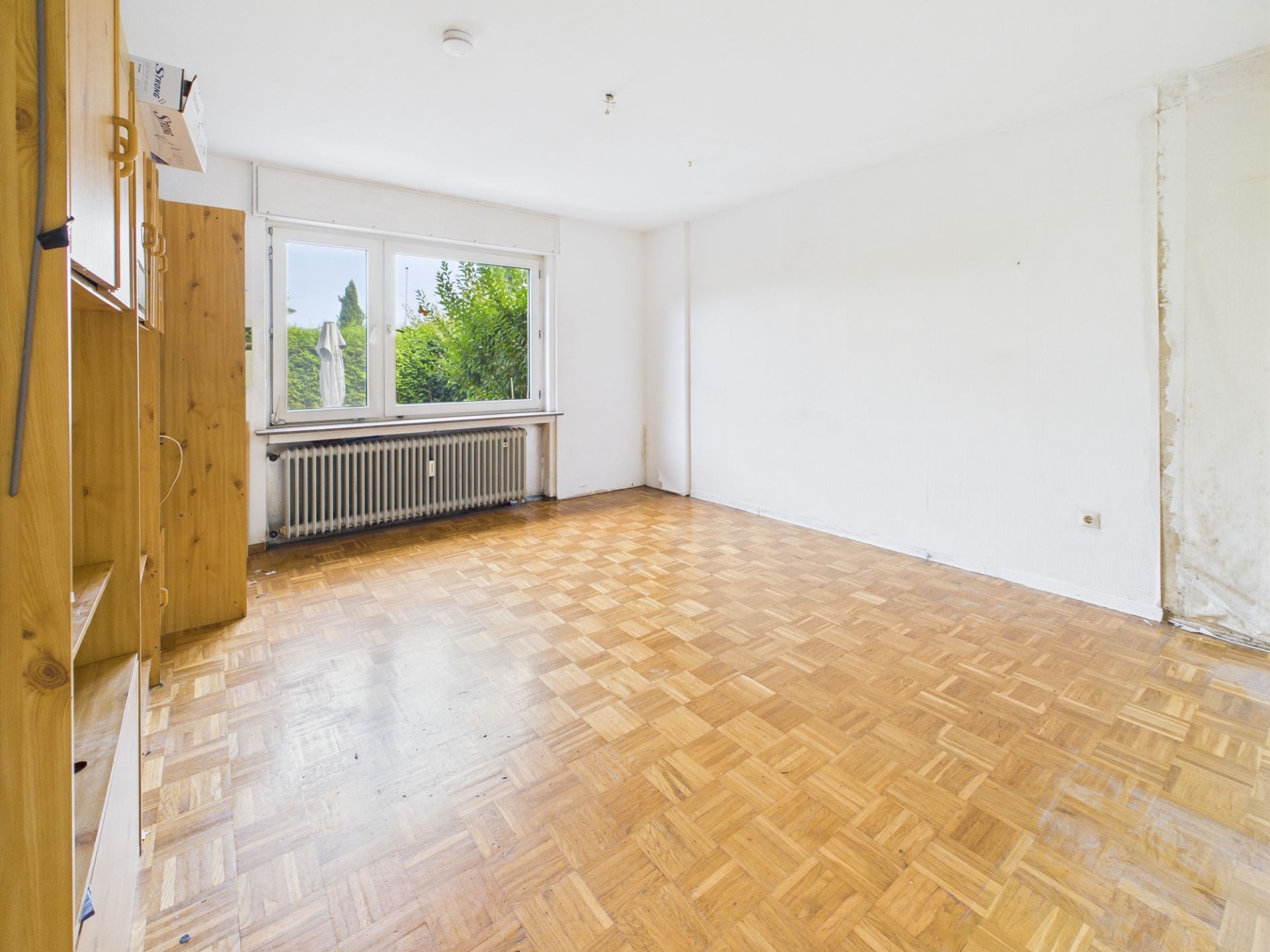 Helles, leeres Zimmer mit Parkettboden, großen Fenstern und einem Heizkörper unter dem Fenster.