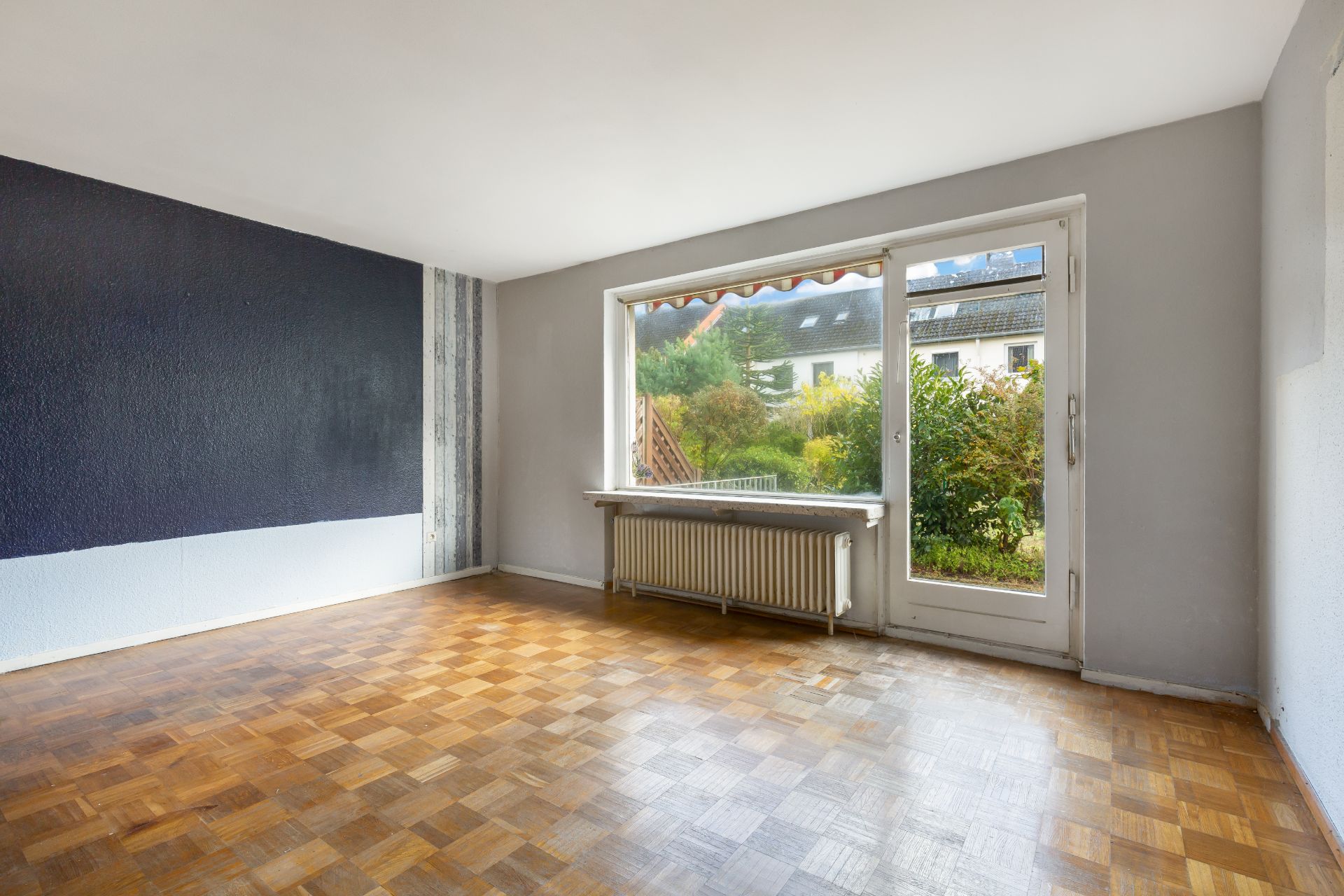Leeres Wohnzimmer mit Parkettboden, großer Fensterfront und Tür zum Garten.
