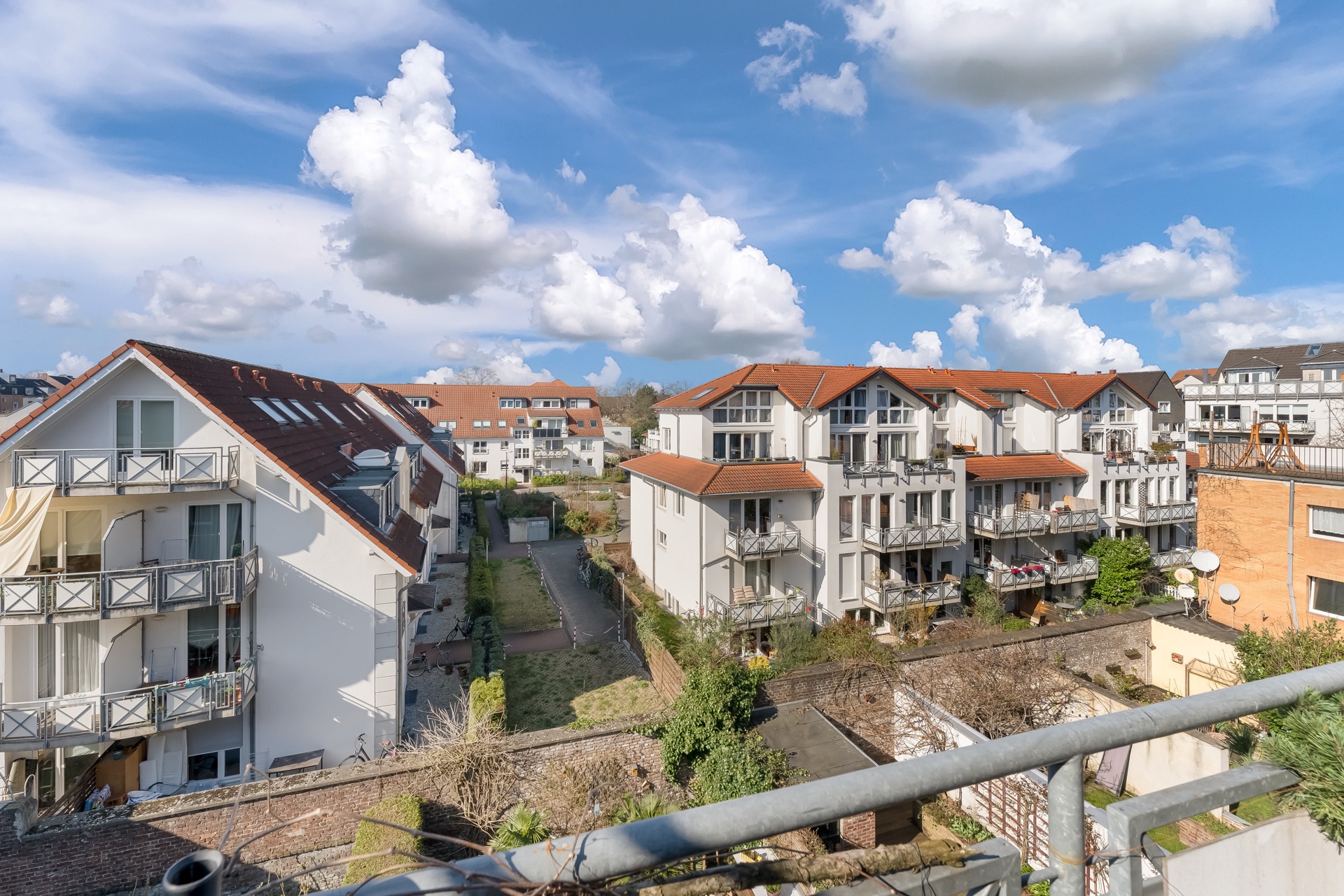 Mehrere moderne Wohnhäuser mit Balkonen, rotem Dach und Gartenbereichen bei sonnigem Wetter.