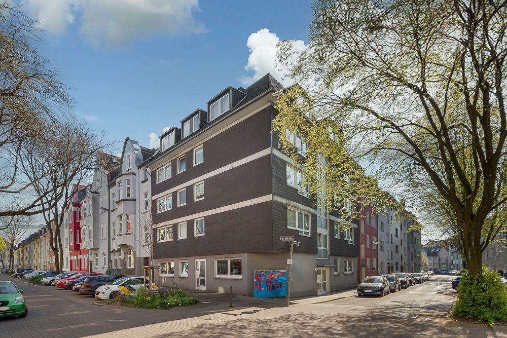 Mehrfamilienhaus an einer Straßenecke mit geparkten Autos und Bäumen im Vordergrund.
