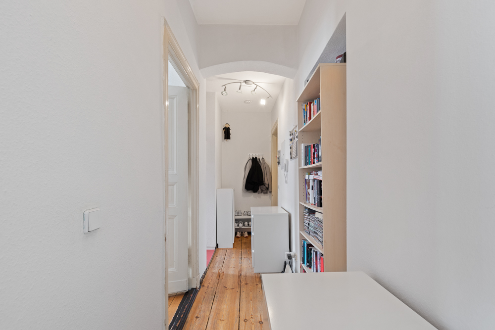 Heller Flur mit Holzboden, Bücherregal und Garderobe mit Jacken am Ende des Gangs.