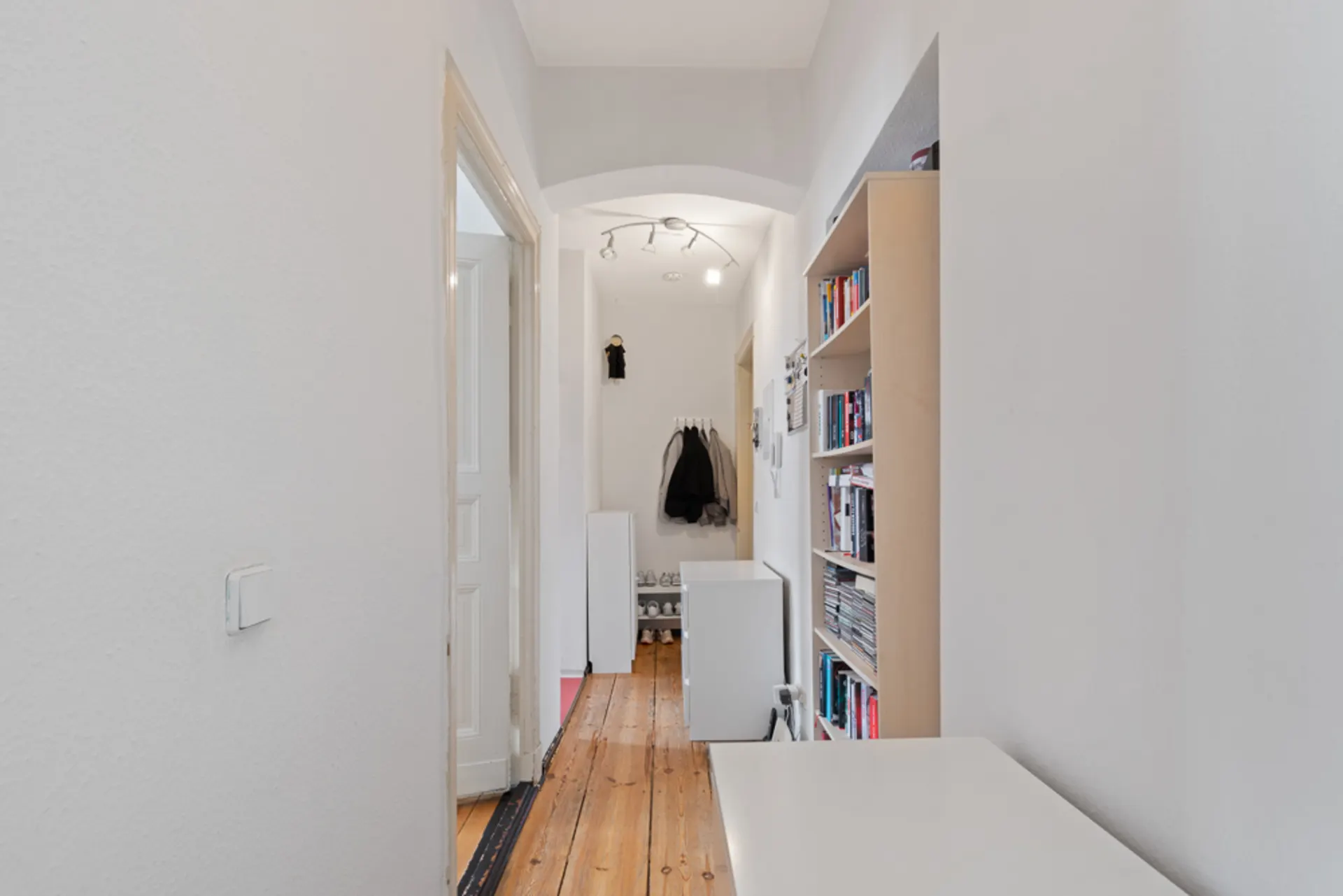 Heller Flur mit Holzboden, Bücherregal und Garderobe mit Jacken am Ende des Gangs.
