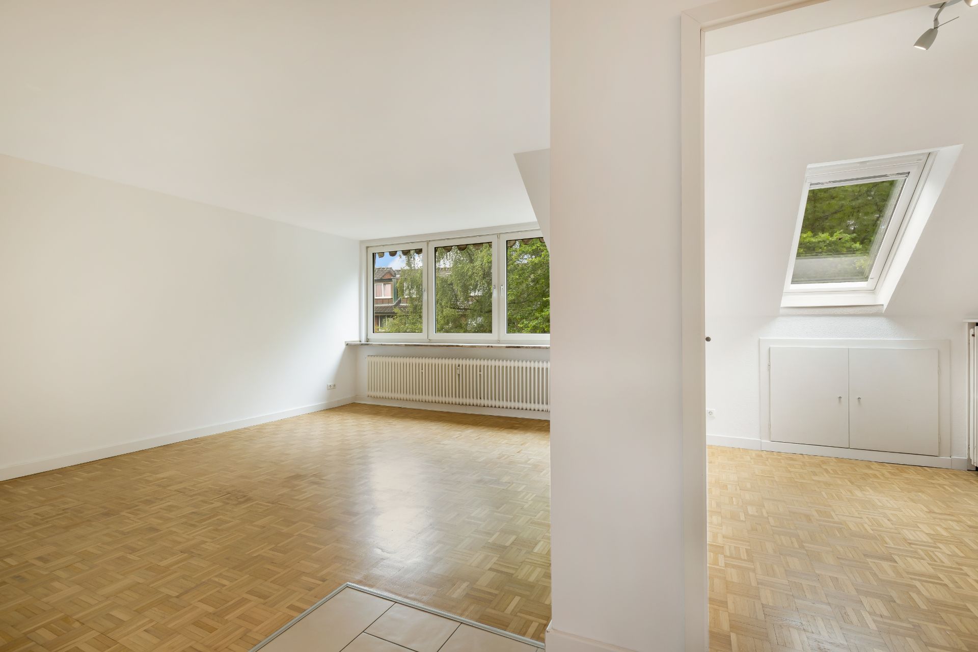 Helles Zimmer mit Parkettboden und großen Fenstern, angrenzend ein weiteres Zimmer mit Dachfenster.