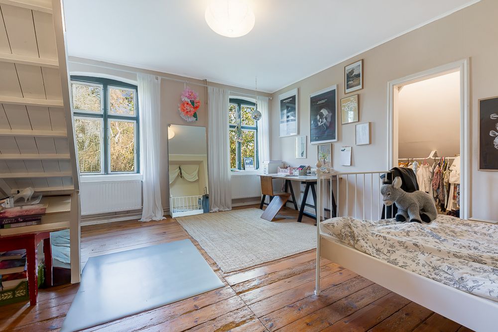 Helles Schlafzimmer mit Holzboden, Bett, Schreibtisch, Spiegel und großen Fenstern.