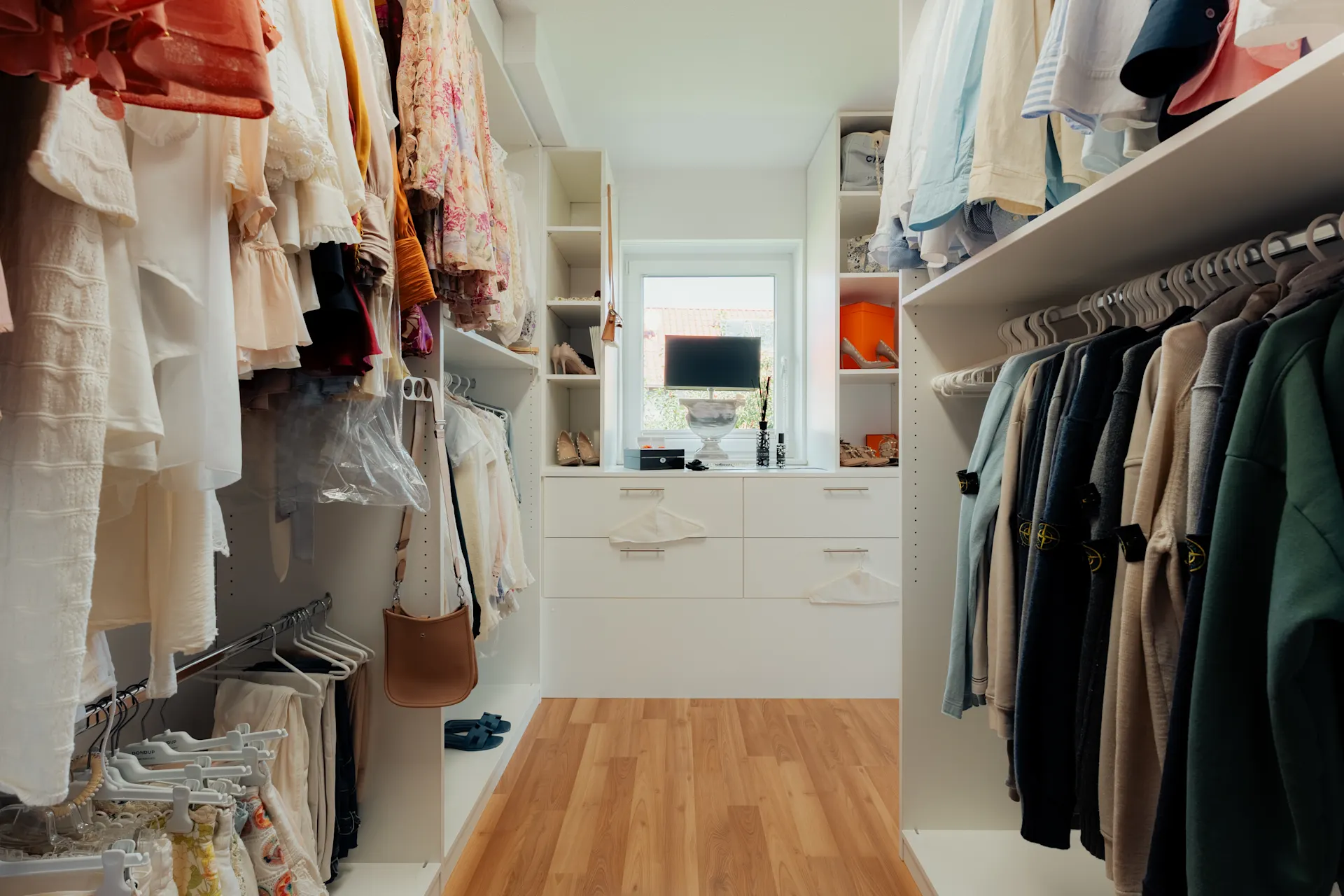Geräumiger begehbarer Kleiderschrank mit Kleidung, Regalen, Schubladen und einem Fenster im Hintergrund.