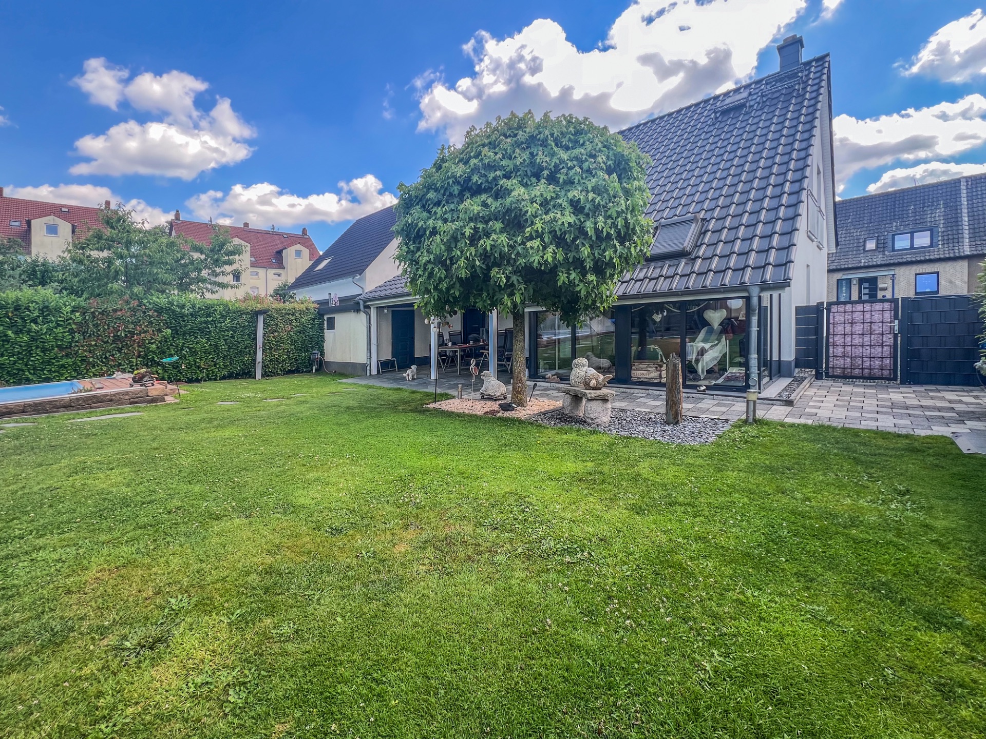 Modernes Einfamilienhaus mit großem Garten, Baum in der Mitte und Terrasse unter blauem Himmel.