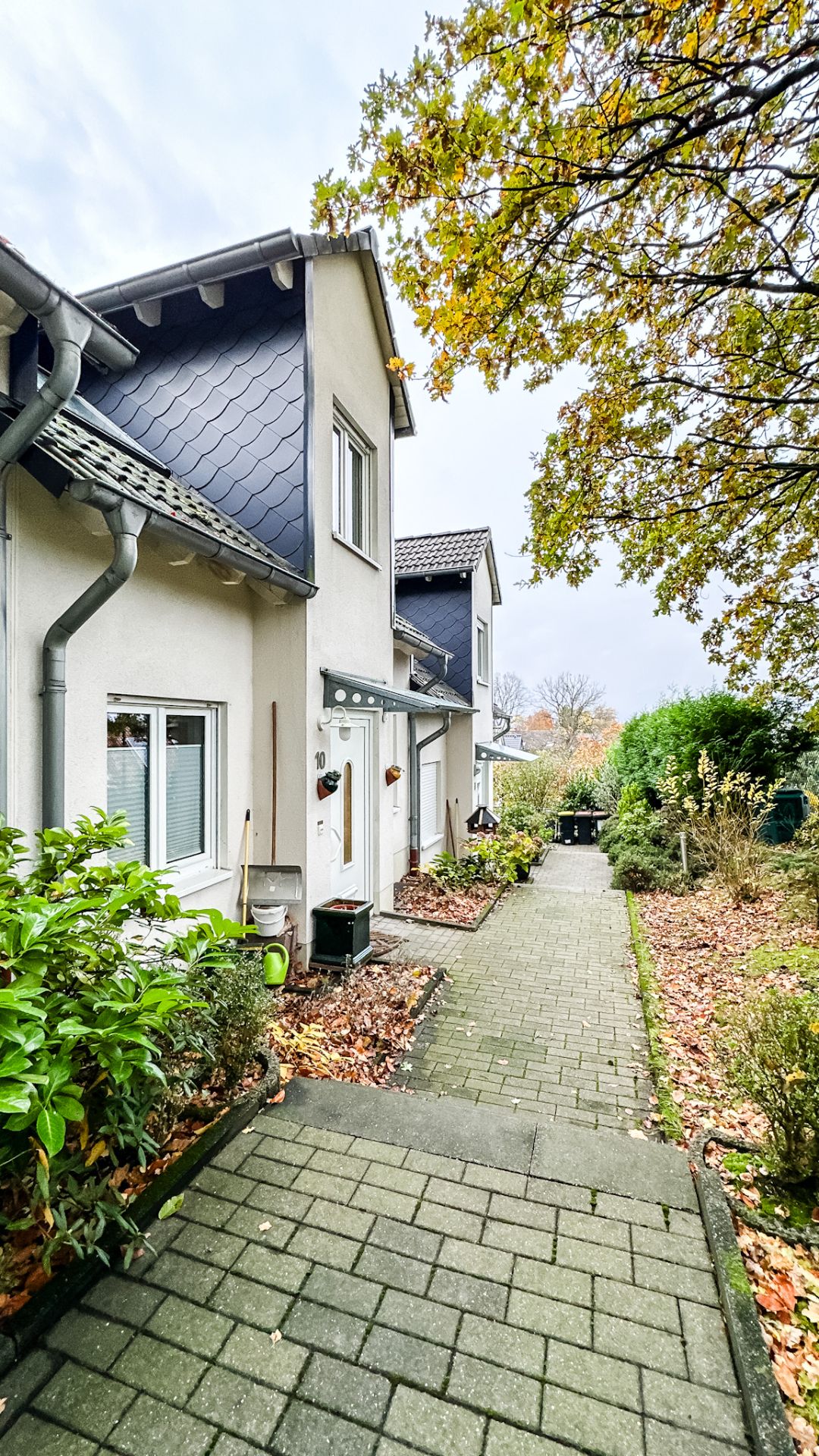 Gepflasterter Weg vor einem Reihenhaus mit kleinen Gärten und herbstlichen Bäumen.