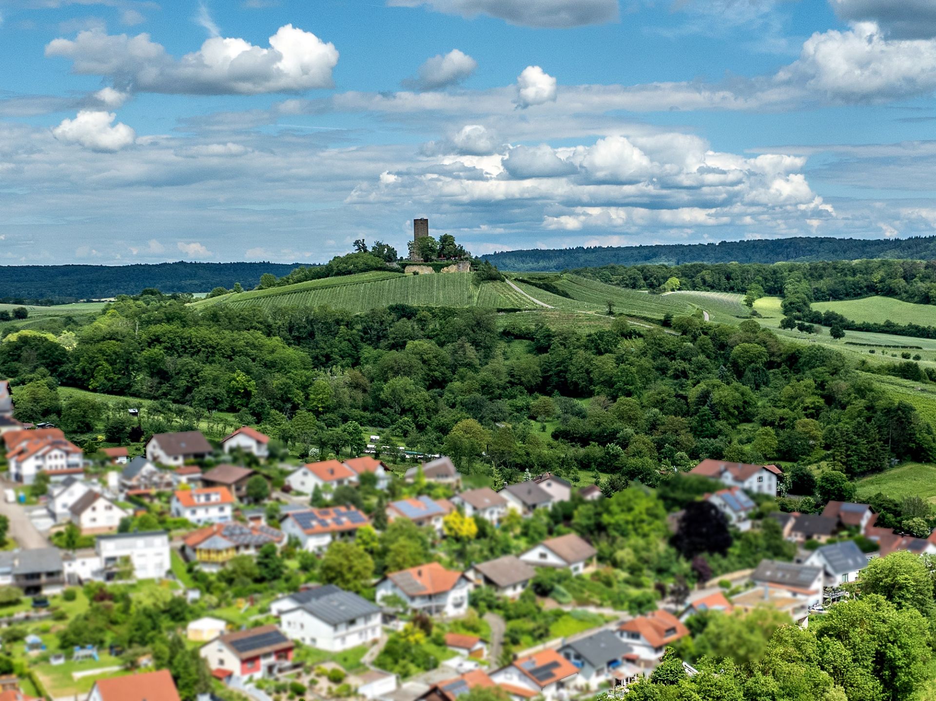 Blick auf ein Wohngebiet mit Häusern, dahinter bewaldete Hügel und eine Burg auf dem Hügel.