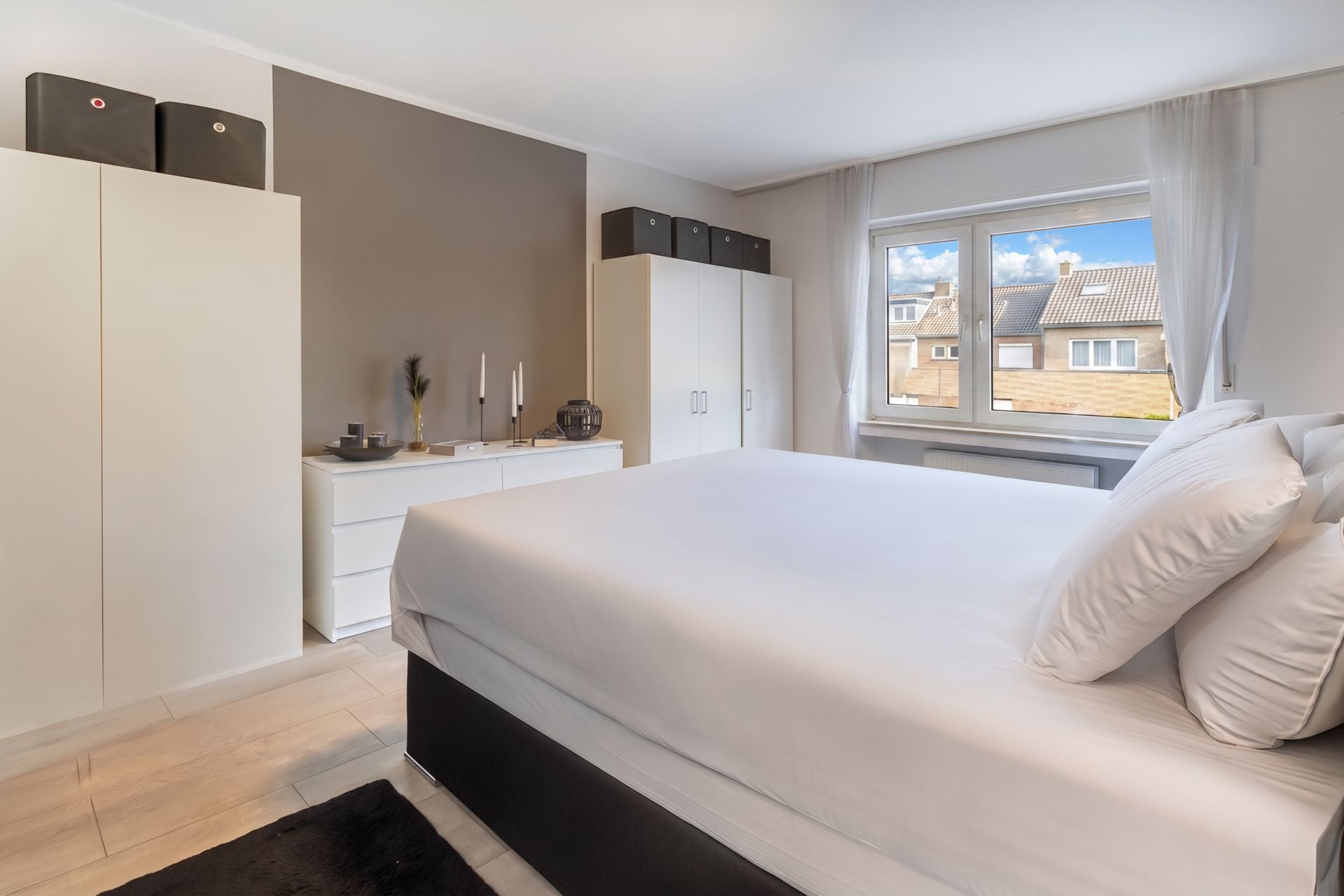 Helles Schlafzimmer mit großem Bett, weißen Schränken, Kommode und Fenster mit Blick auf Nachbarhäuser.