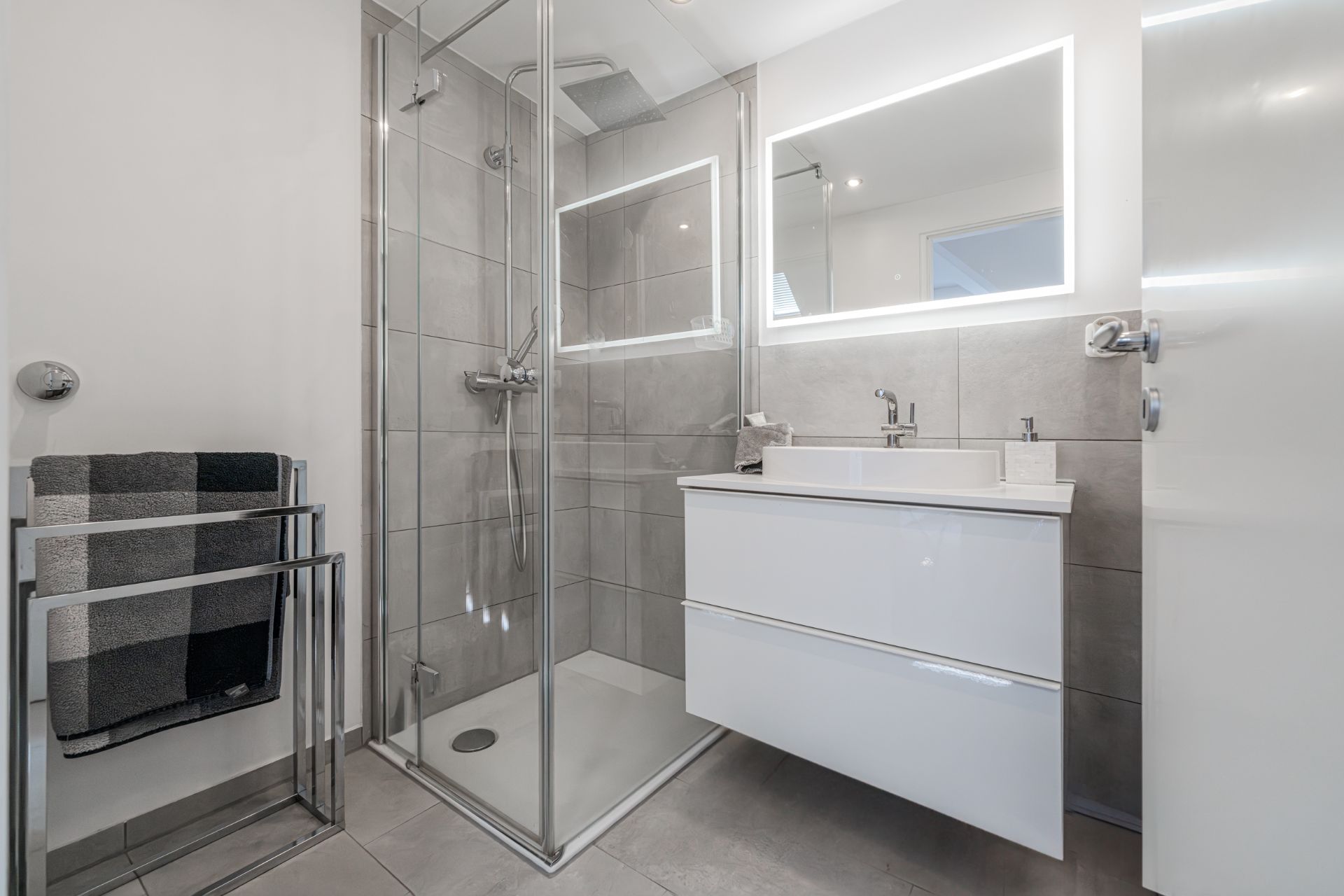 Modernes Bad mit Glasdusche, weißem Waschbecken, beleuchtetem Spiegel und Handtuchhalter.