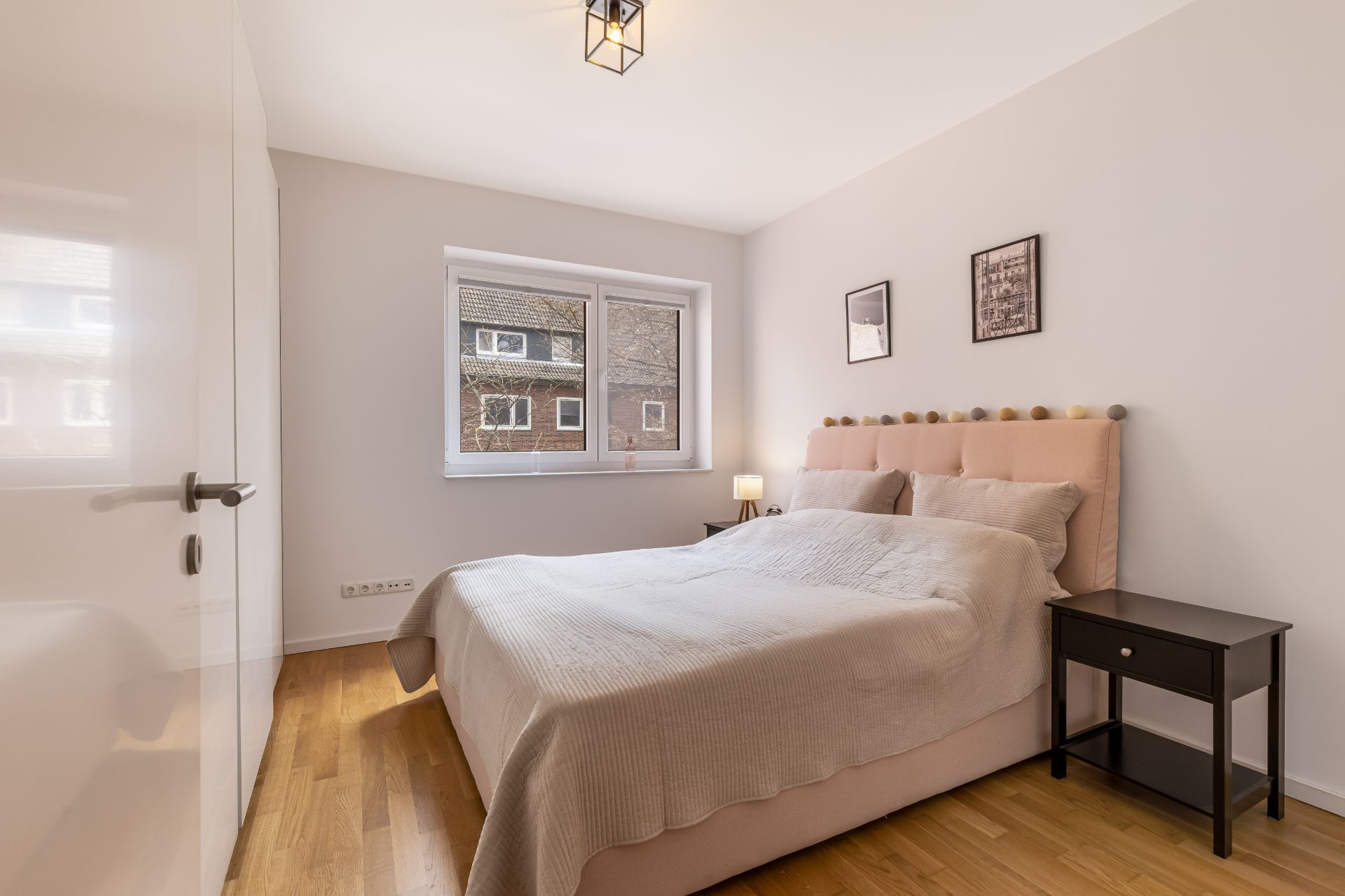 Helles Schlafzimmer mit Doppelbett, zwei Nachttischen und Fenster zu einer Wohnhausansicht.