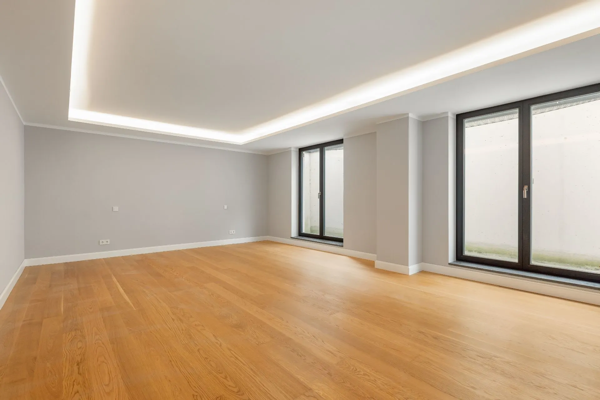 Leerer Raum mit Holzfußboden, großen Fenstern und weiß gestrichenen Wänden.