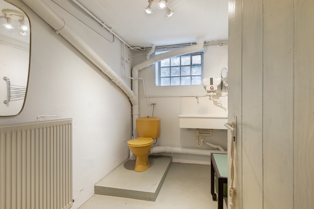 Kleines Badezimmer mit gelber Toilette, Waschbecken, Spiegel und Heizkörper, Kellerfenster im Hintergrund.