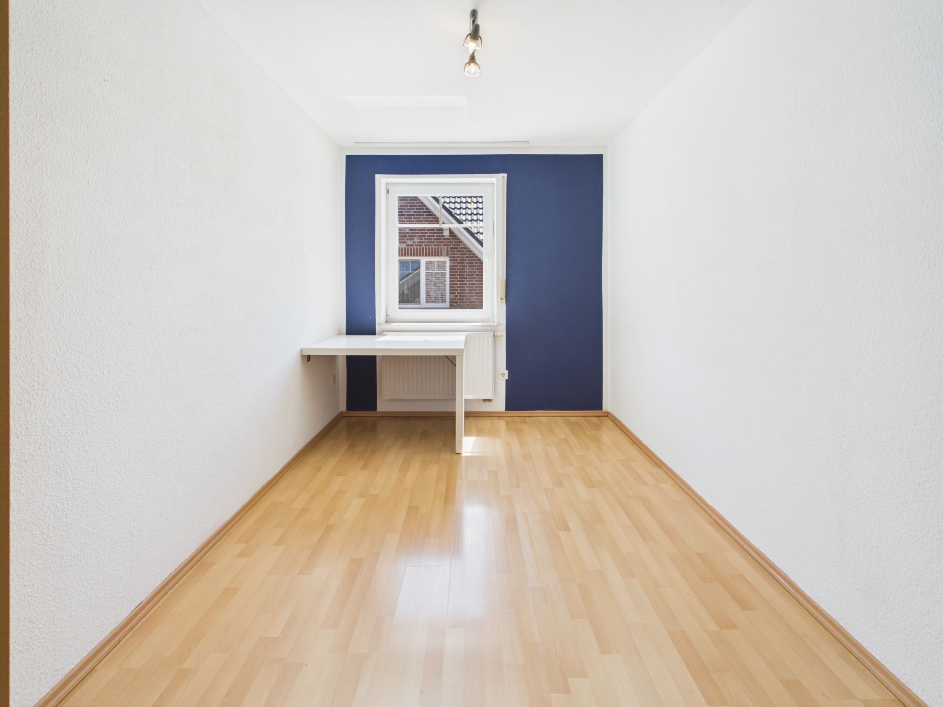 Helles Zimmer mit Holzfußboden, blau-weißer Wand, Fenster und kleinem Wandtisch.