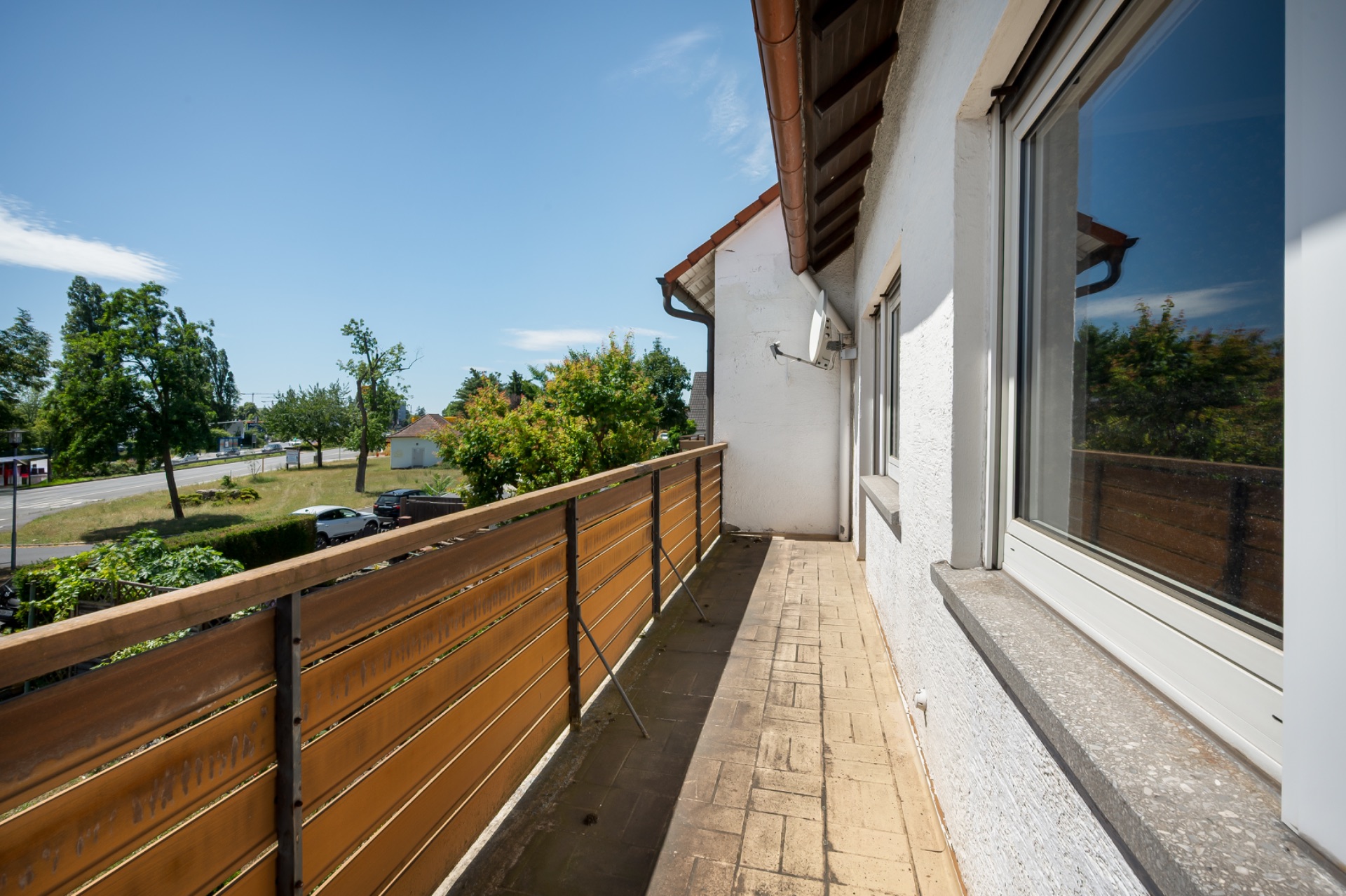 Langer Balkon mit braunem Geländer an einem weißen Wohnhaus, Blick auf Bäume und Straße.