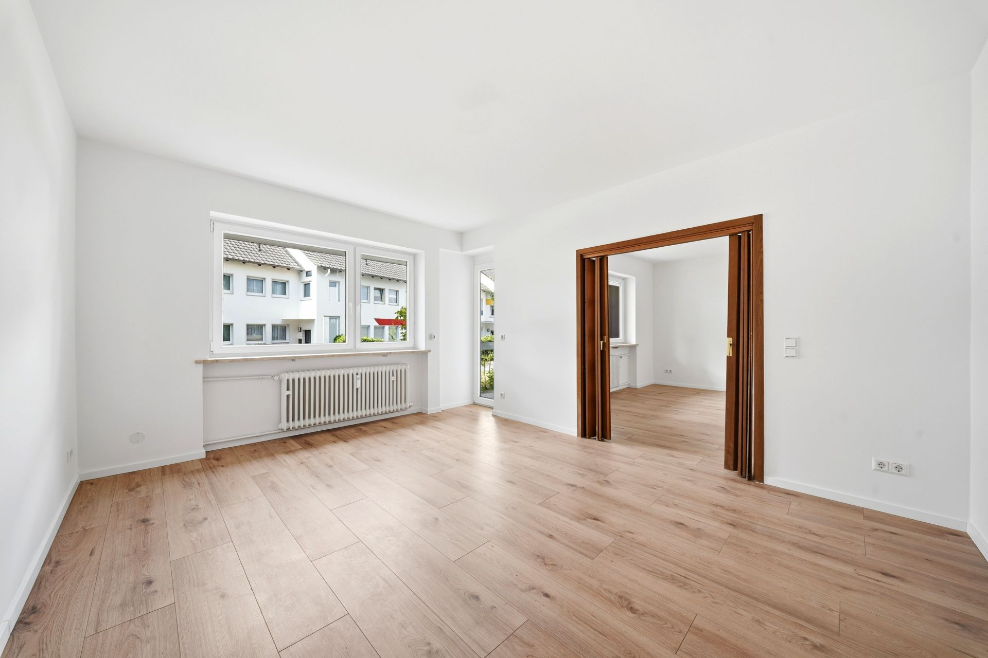 Helles Wohnzimmer mit Holzfußboden, großem Fenster und Durchgang in einen weiteren Raum.