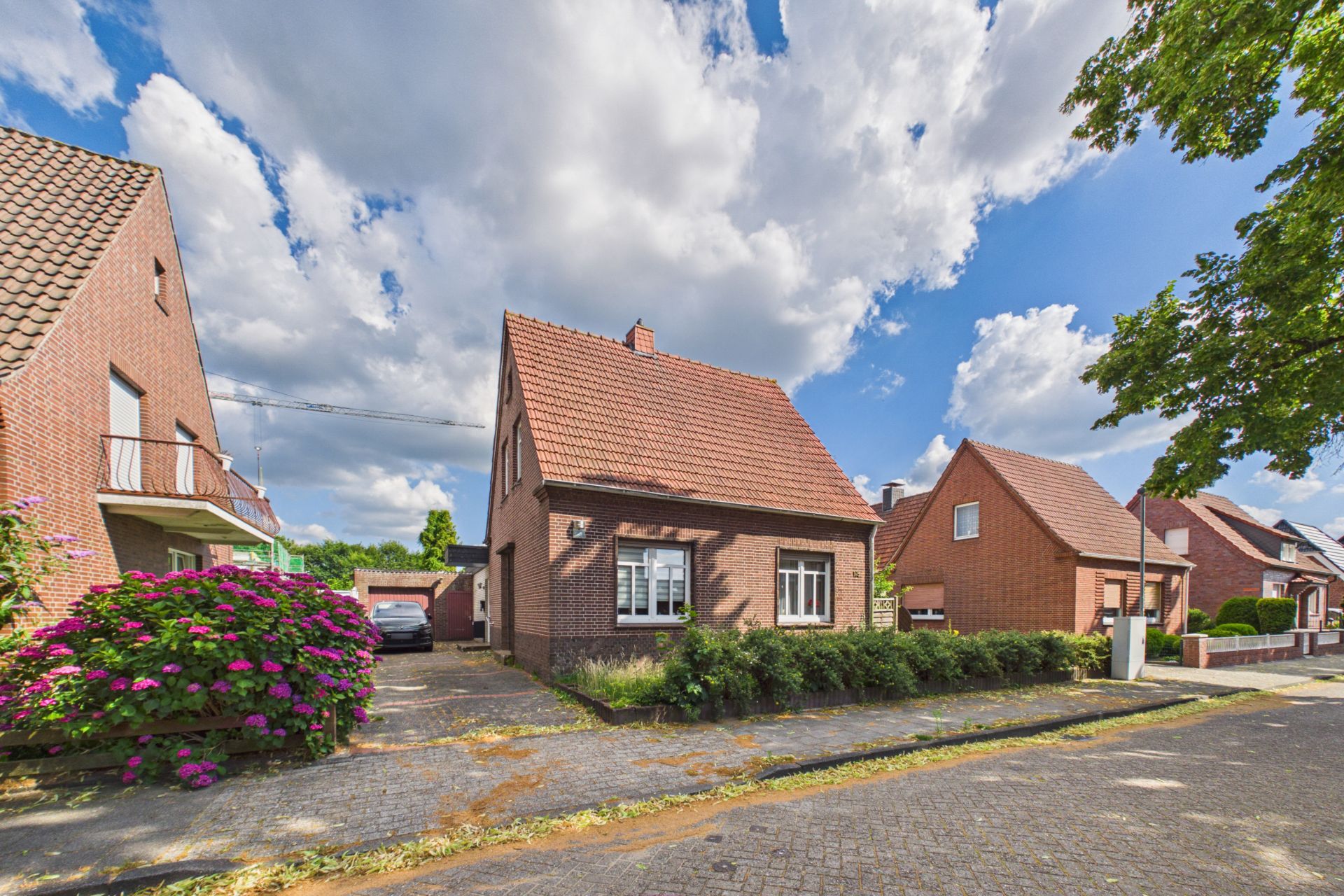 Rotes Einfamilienhaus mit Satteldach, Vorgarten und bepflanzter Einfahrt in einer Wohnsiedlung.