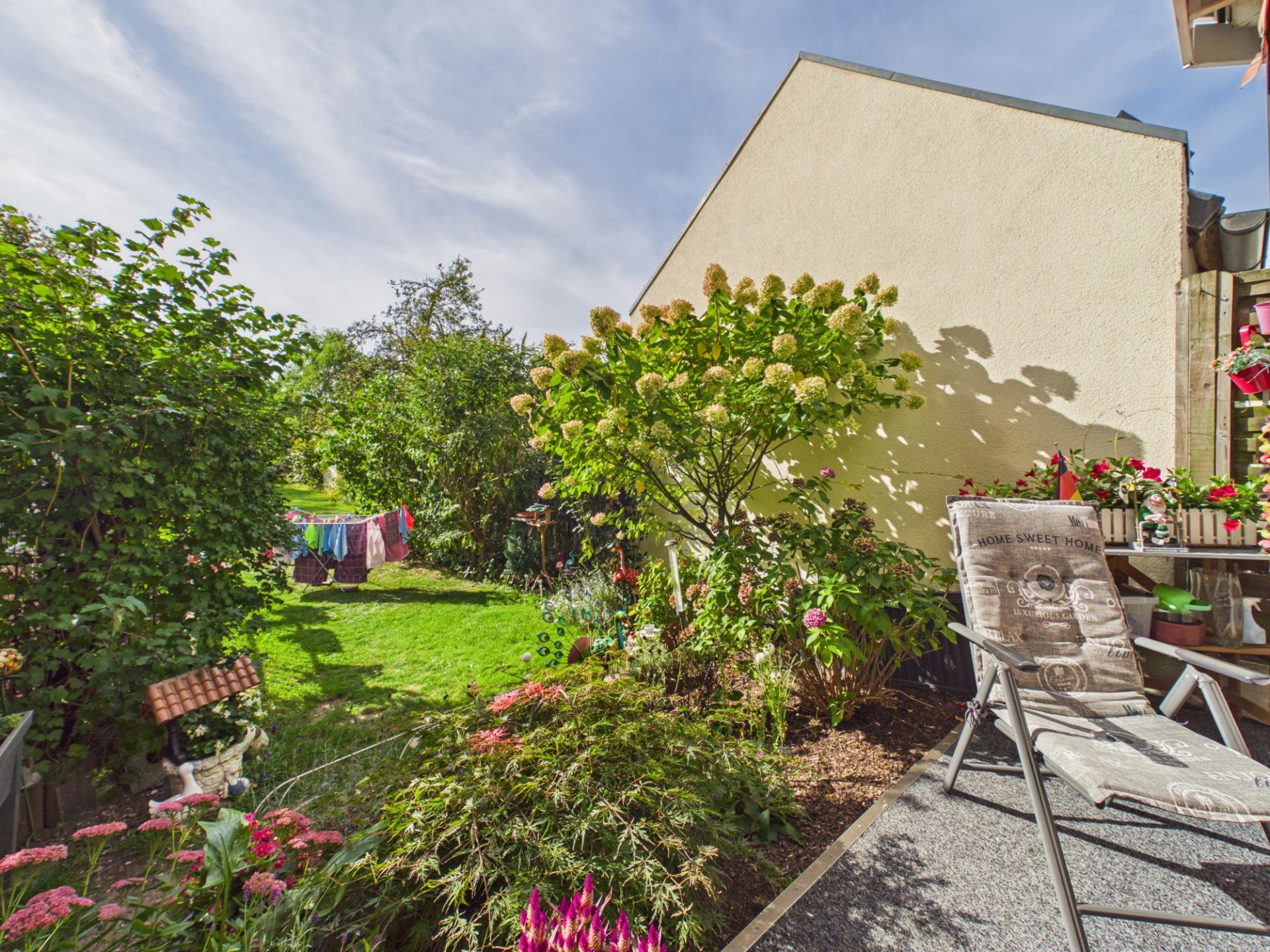 Gepflegter Garten mit Blumen, Sträuchern, Liegestuhl und Wäscheleine im Hintergrund.