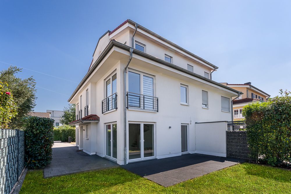 Modernes, weißes Wohnhaus mit Balkon und Garten, umgeben von Hecke und Zaun.