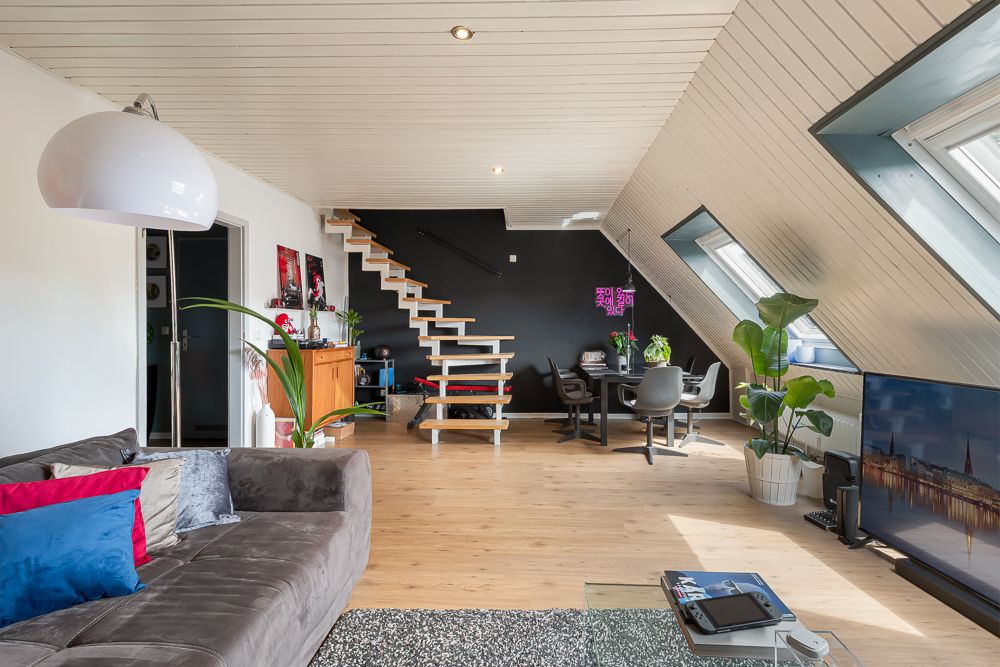 Modernes Wohnzimmer mit Sofa, offenem Treppenaufgang, Esstisch und großen Fenstern unter Dachschräge.