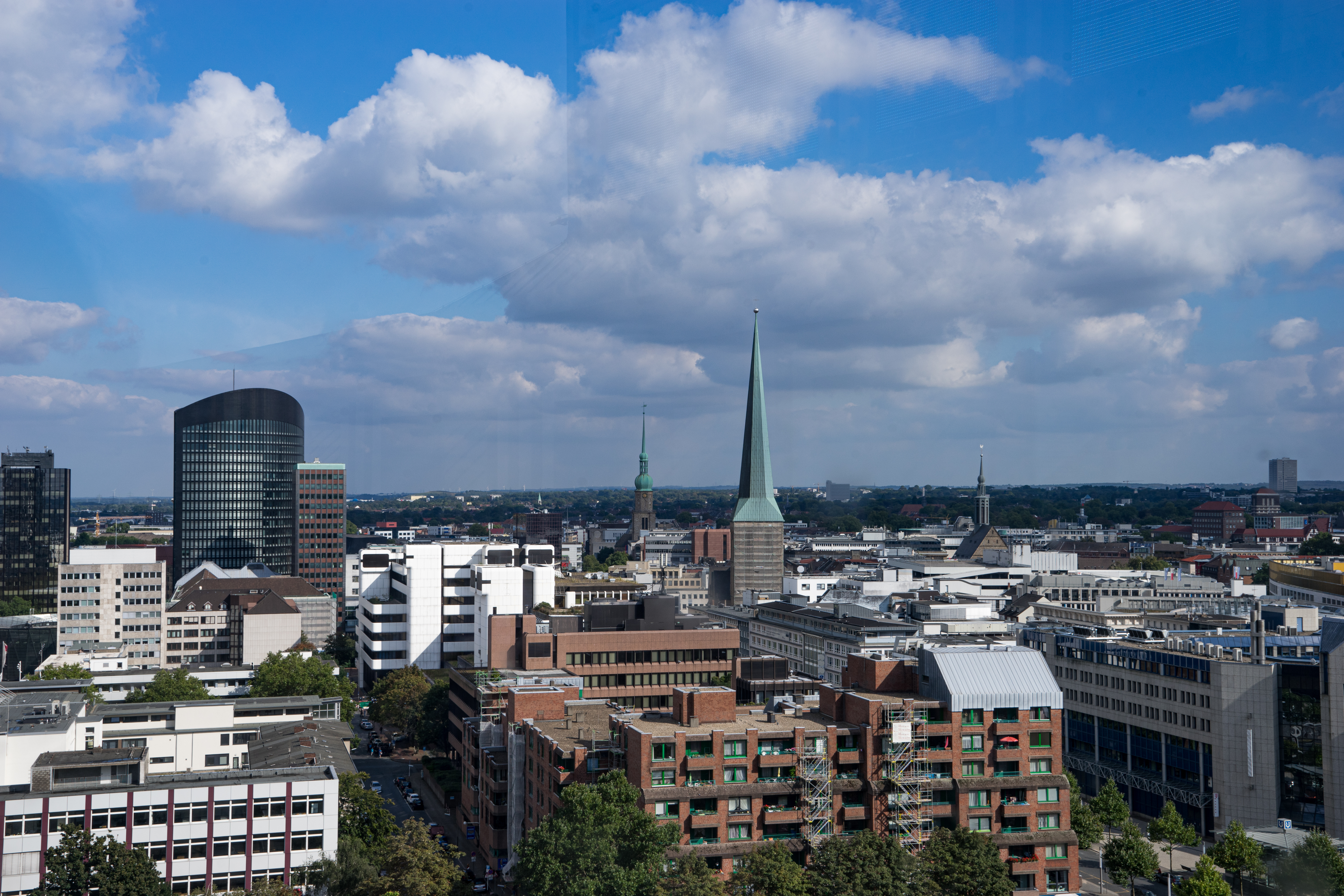 Stadtansicht mit modernen und historischen Gebäuden, mehrere Kirchtürme, teilweise bewölkter Himmel.