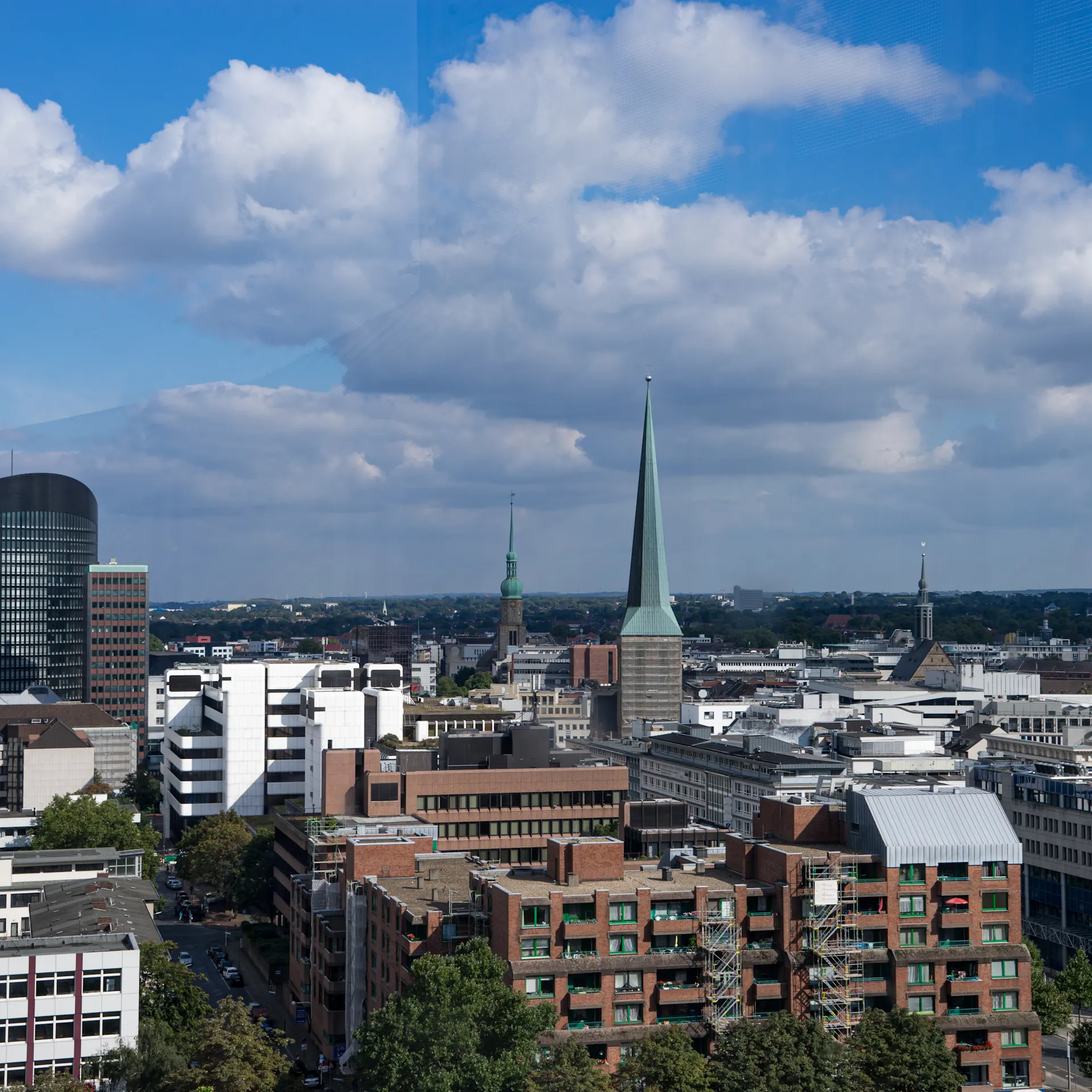 Stadtansicht mit modernen und historischen Gebäuden, mehrere Kirchtürme, teilweise bewölkter Himmel.