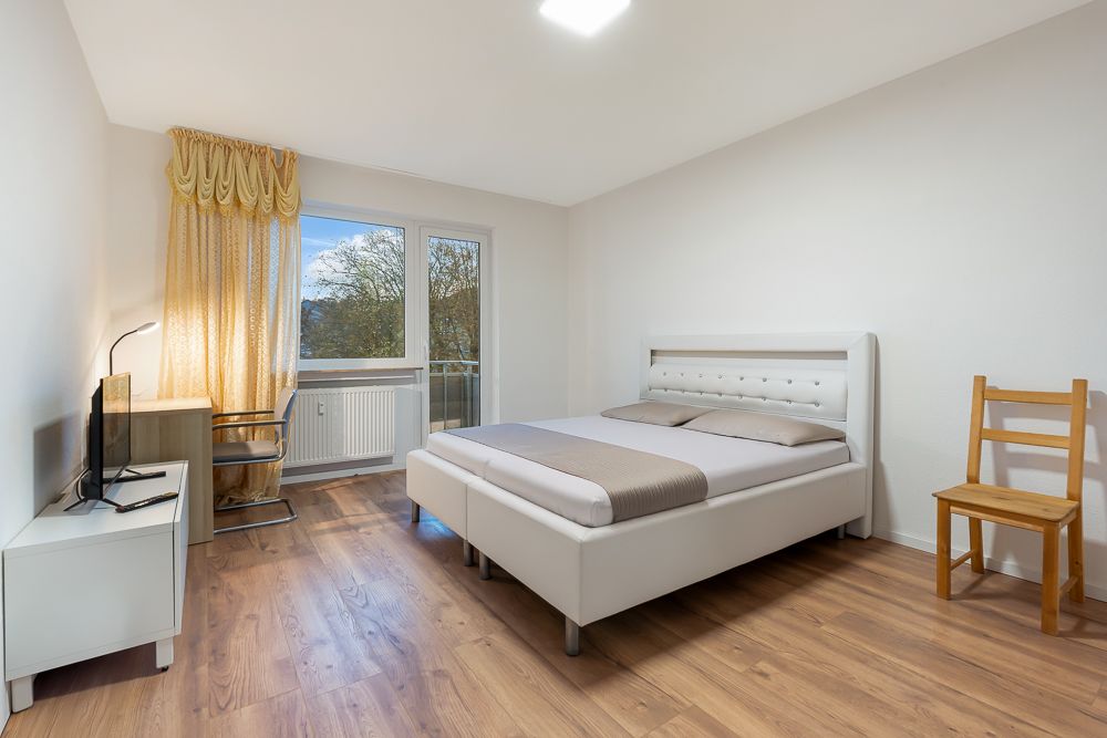 Helles Schlafzimmer mit Doppelbett, Schreibtisch, Stuhl, Fernseher und Fenster mit Vorhängen.
