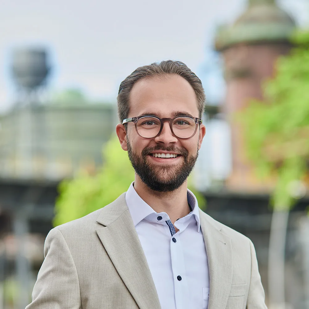 Mann mit Brille und weißem Hemd steht in einem hellen Flur mit Pflanzen im Hintergrund.